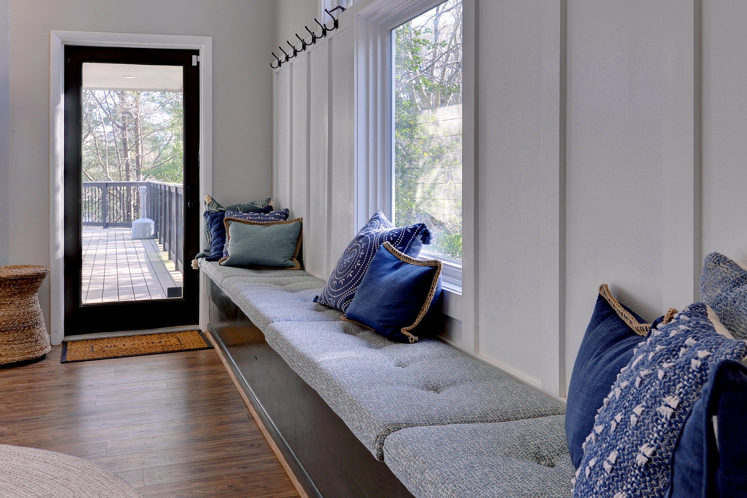 Sunroom with built-in window seat decorated with blue and teal pillows, next to a sliding glass door leading to a wooden deck with a view of trees.