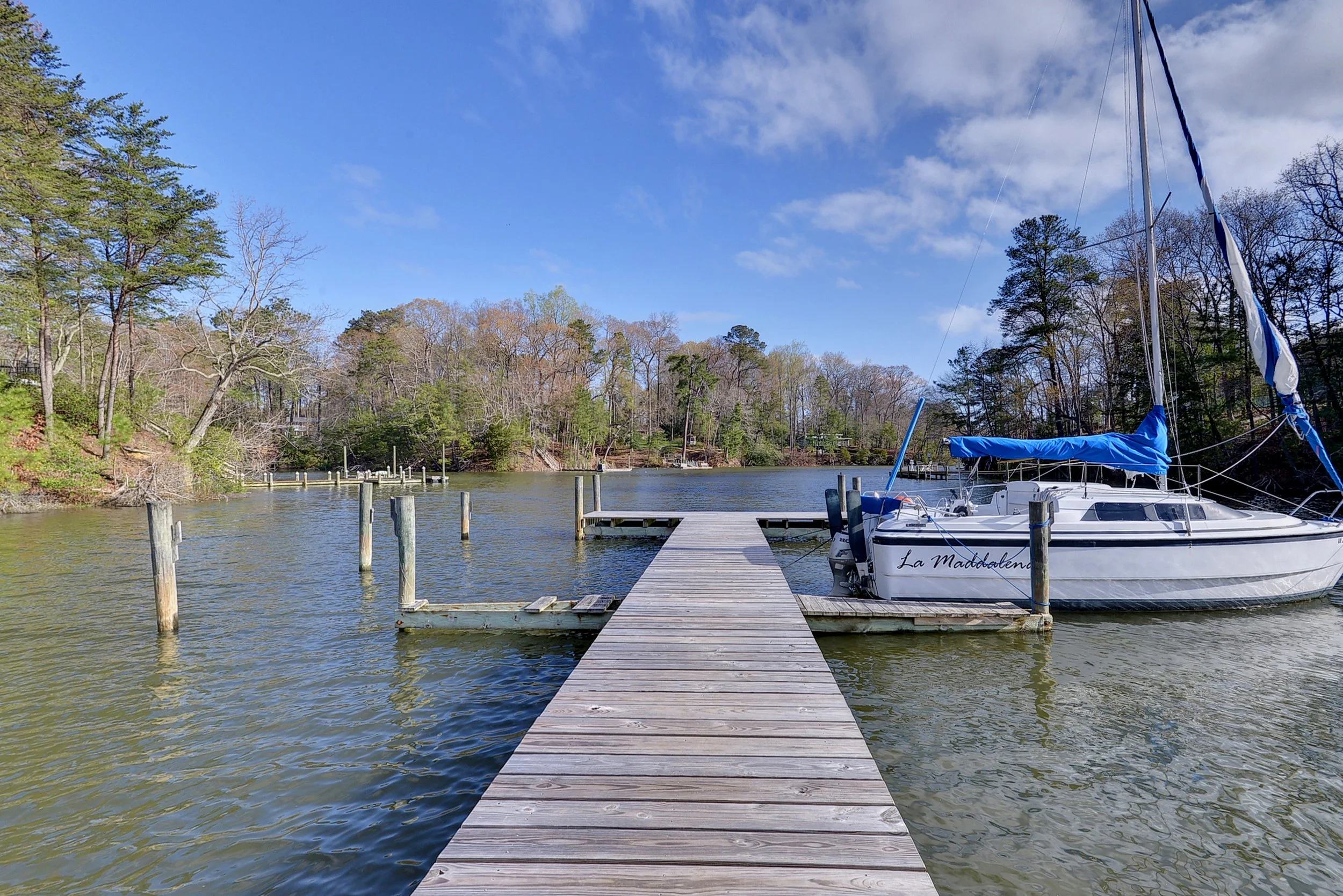 Wooden dock extending into a lake with a sailboat named "La Maddalena" tied to the dock, surrounded by trees and under a partly cloudy sky.
