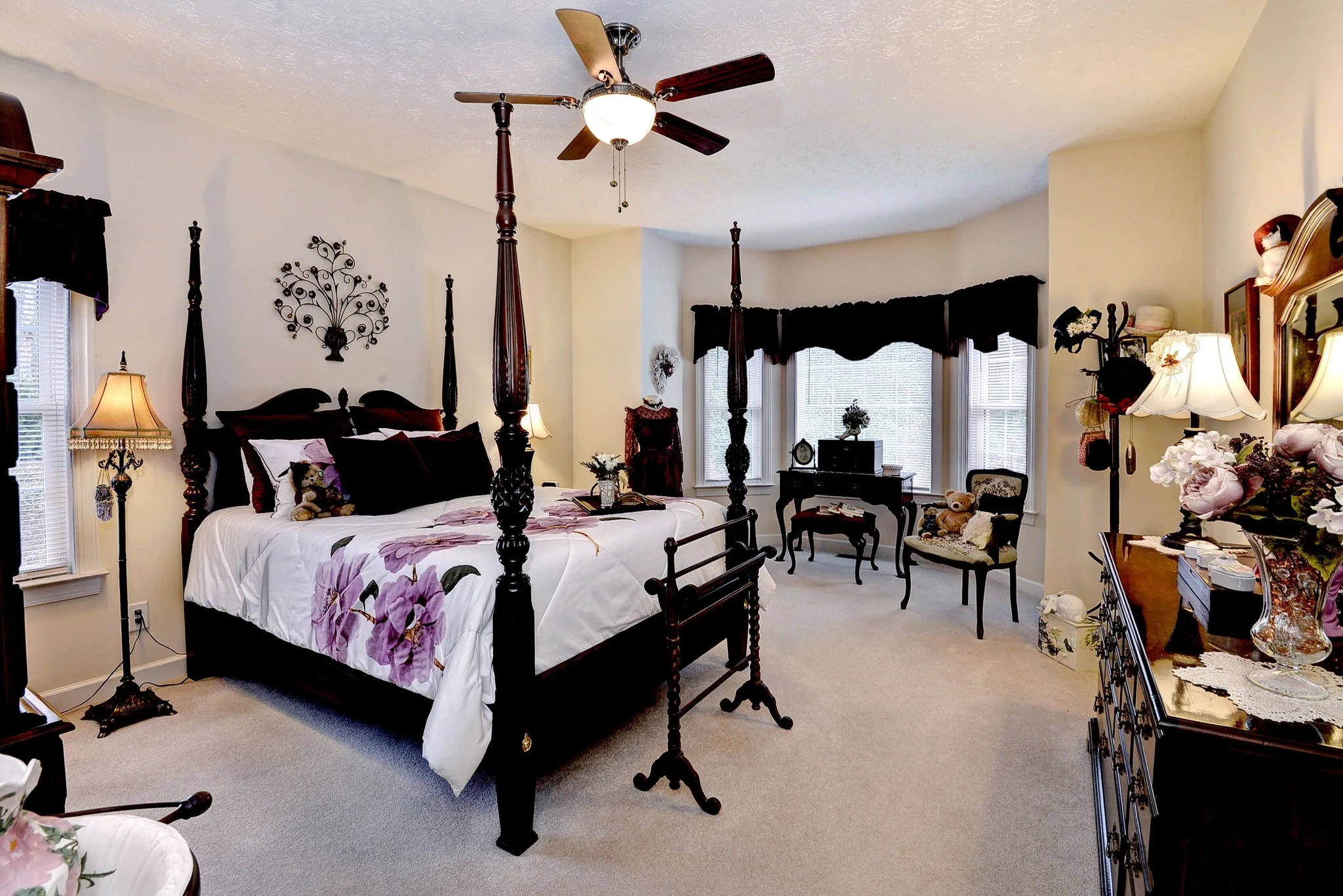 A bedroom with a four-poster bed, floral bedding, and teddy bears. There are lamps, a side table, and vintage furniture, with windows covered by dark curtains.