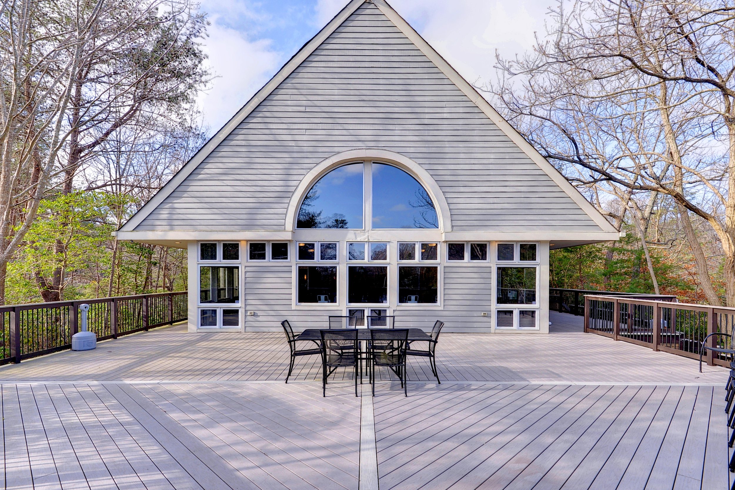 Large wooden deck with black metal patio furniture, surrounded by a railing, in front of a house with gray siding and large windows, set in a wooded area with trees.
