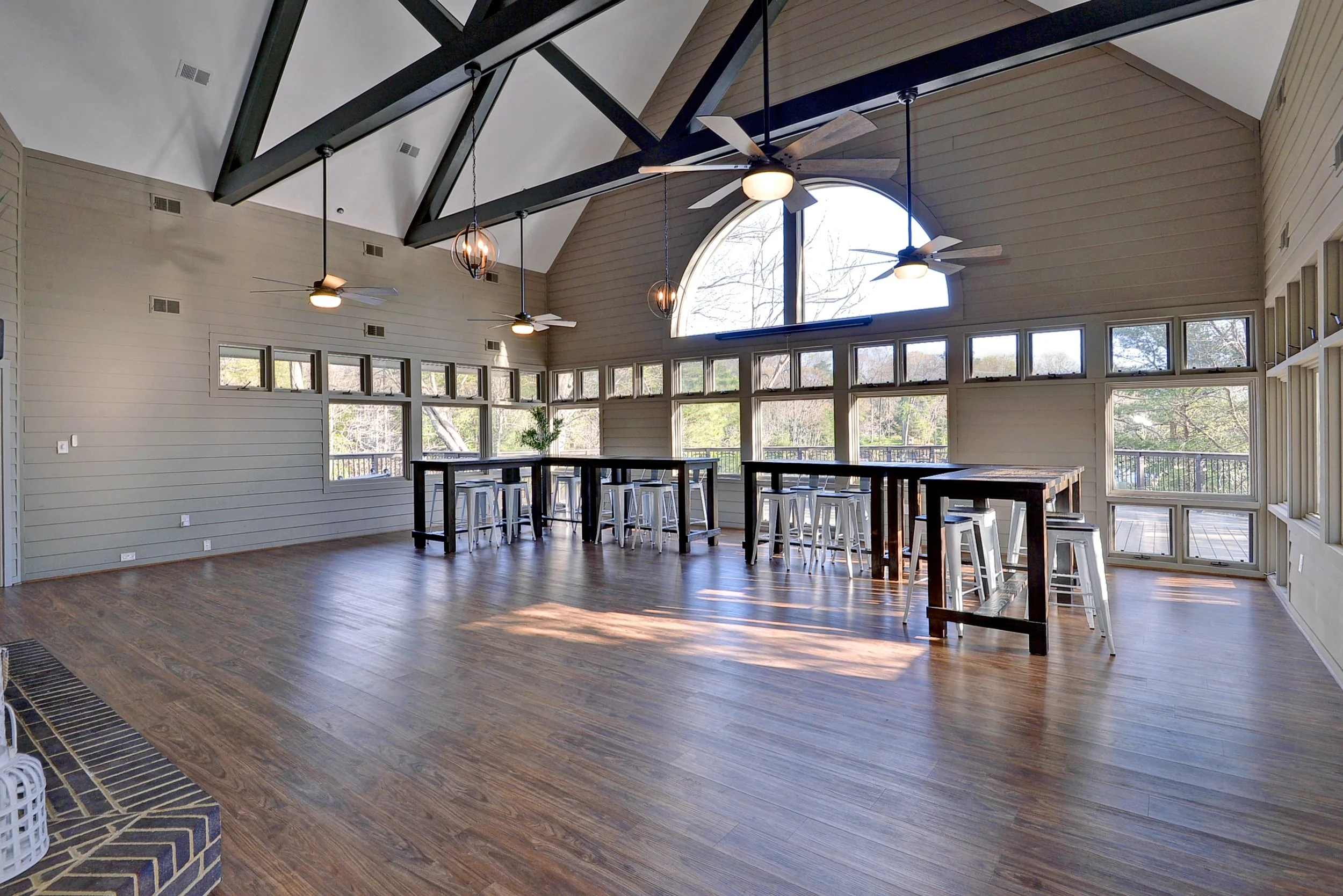 Spacious room with high vaulted ceiling, large windows, wooden floors, ceiling fans, and tall tables with stools, bright with natural light.