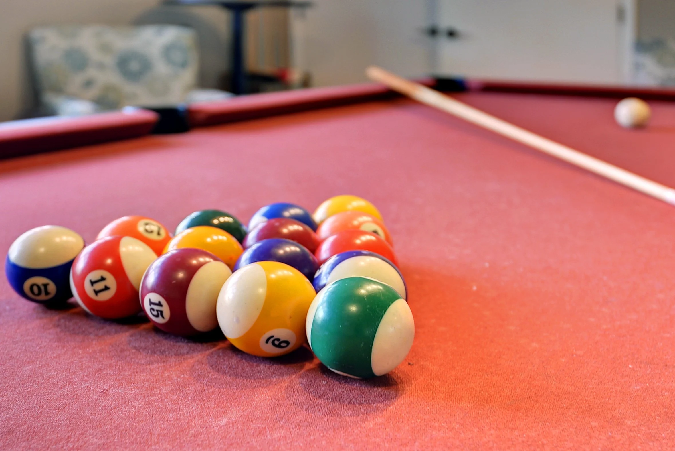 Pool balls grouped together on a red pool table. In the background, the cue stick points toward the white cue ball near the corner pocket.