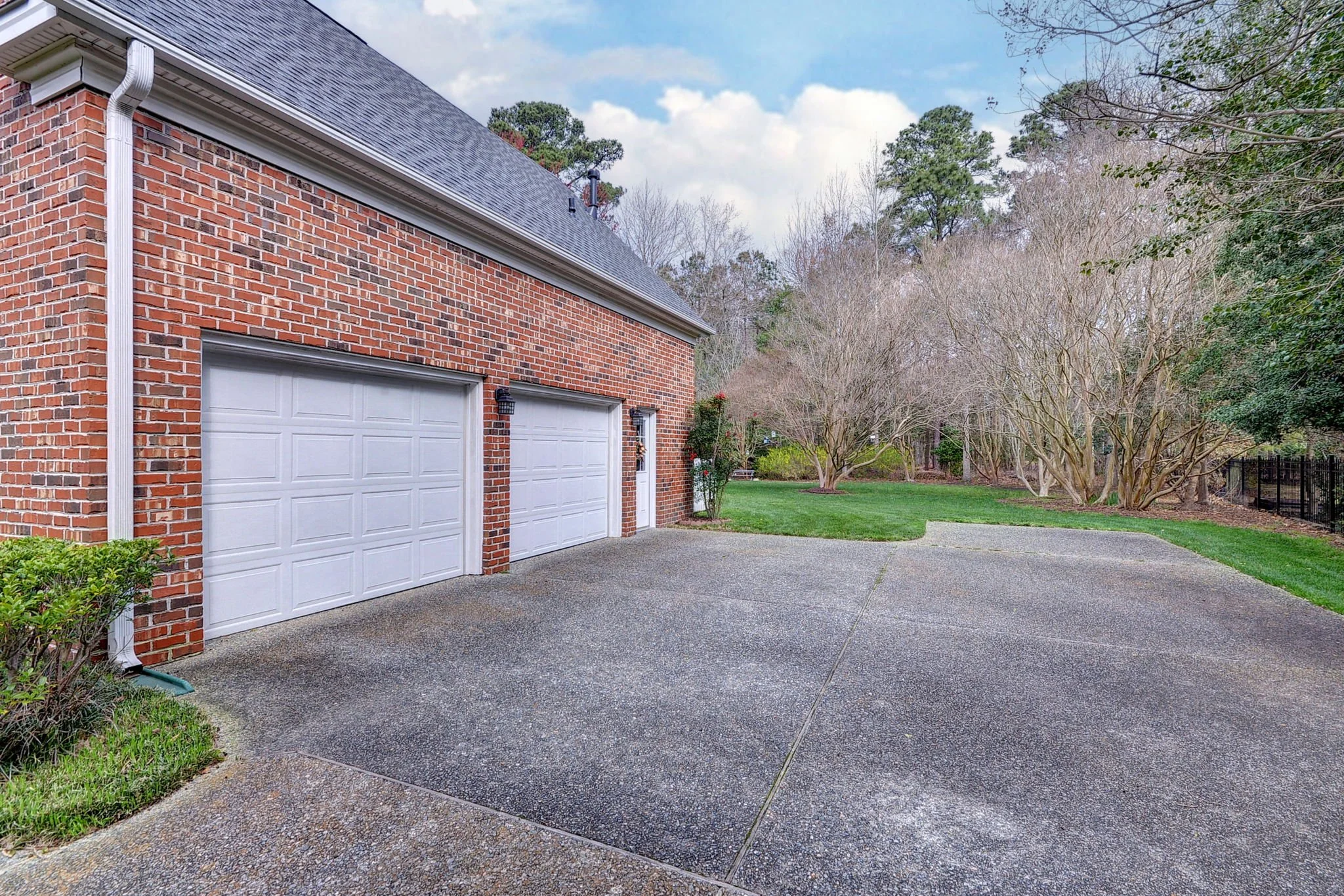 Brick house with two white garage doors and a concrete driveway, surrounded by green grass and trees in a suburban backyard.