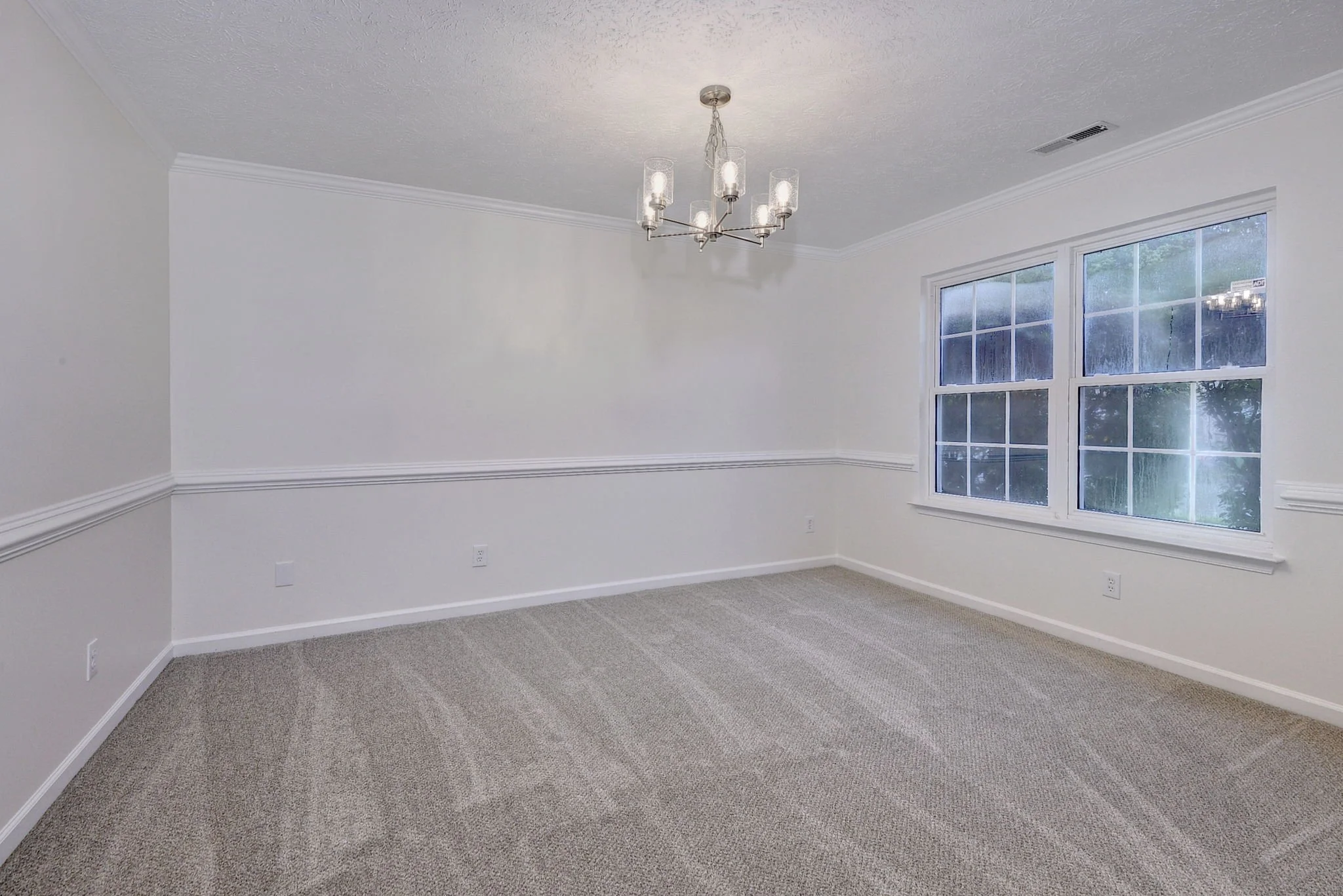 Empty room with beige carpet, white walls with crown molding, large window, and modern chandelier.