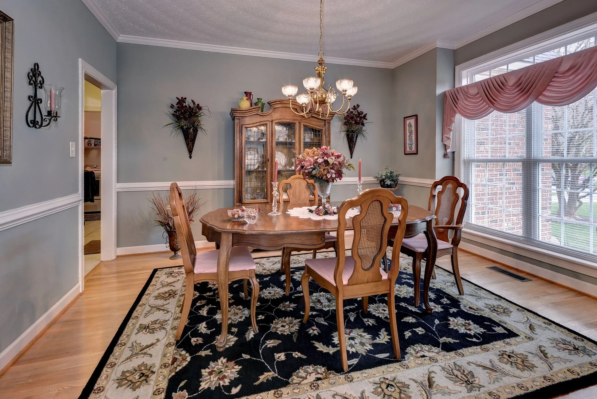 A dining room with a wooden table and six chairs, a chandelier overhead, a china cabinet in the background, a large window with pink drapes, and floral decorations on the walls and table.