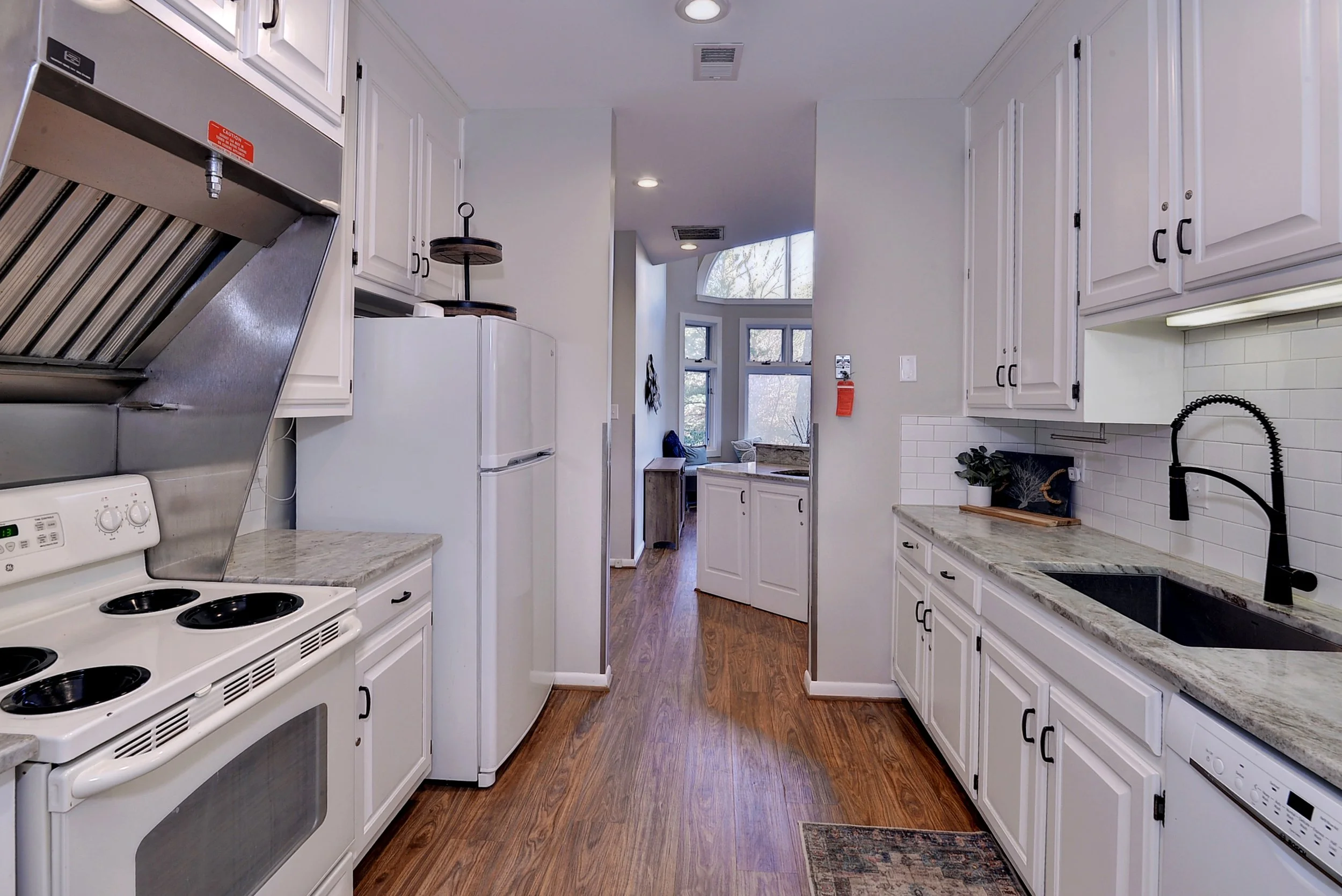 Kitchen with white cabinets, a marble countertop, a black sink, a black faucet, white subway tile backsplash, and a wooden floor leading to a window and sitting area.