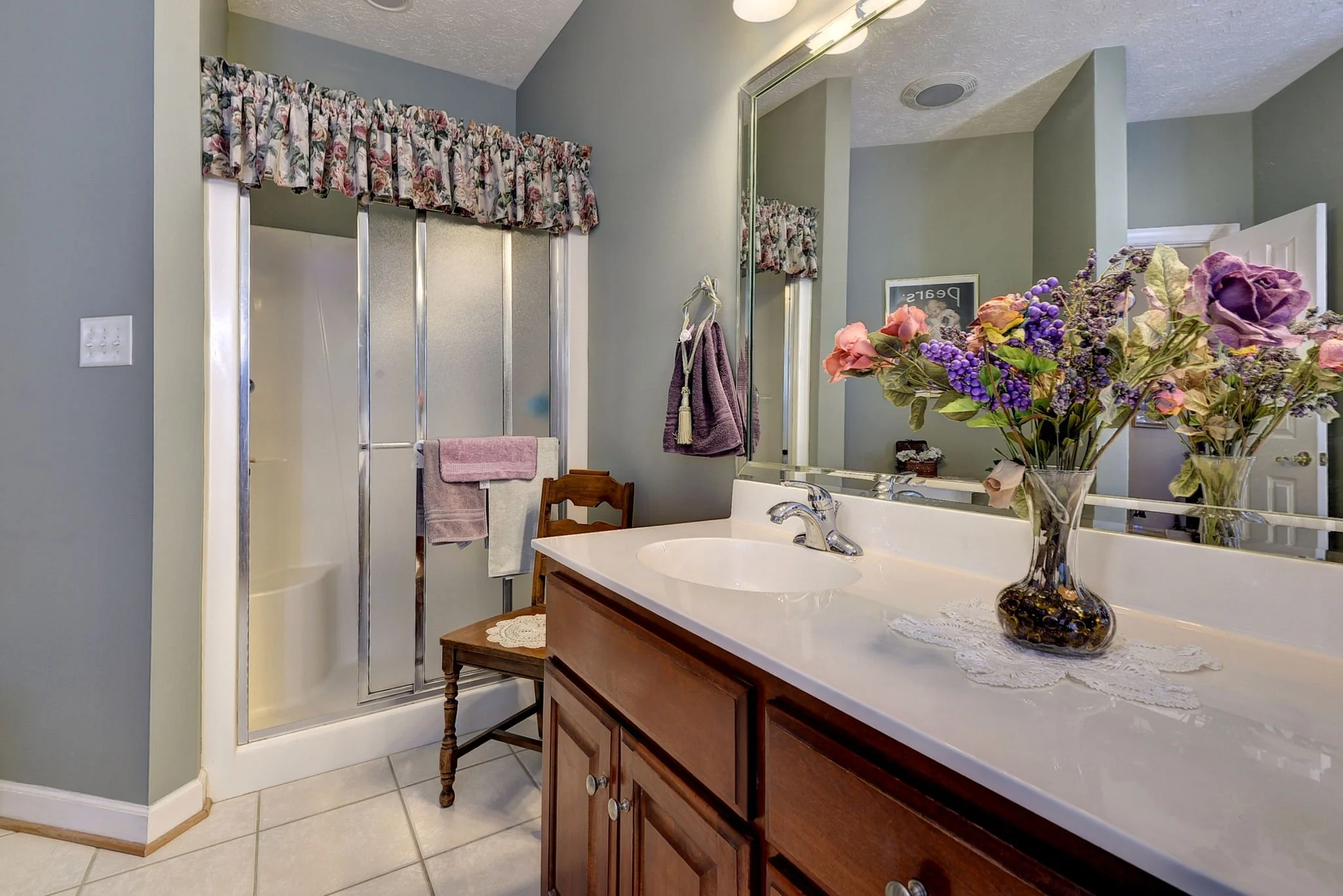 A bathroom with a shower stall, a wooden vanity with a white countertop, and a large mirror. There are pink and purple towels hanging, a vase with pink and purple flowers on the counter, and a small wooden chair with a lace doily.