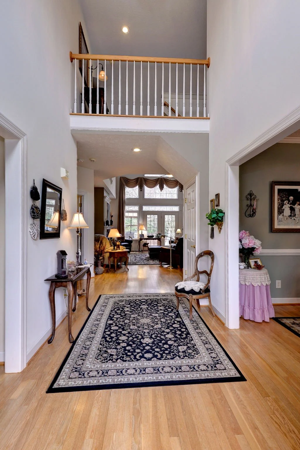 Interior view of a home's entryway with a wooden floor, black and white patterned rug, and white walls. In the background, a living room with large windows, curtains, and sofas is visible. There's a small decorative table with pink flowers on the rig
