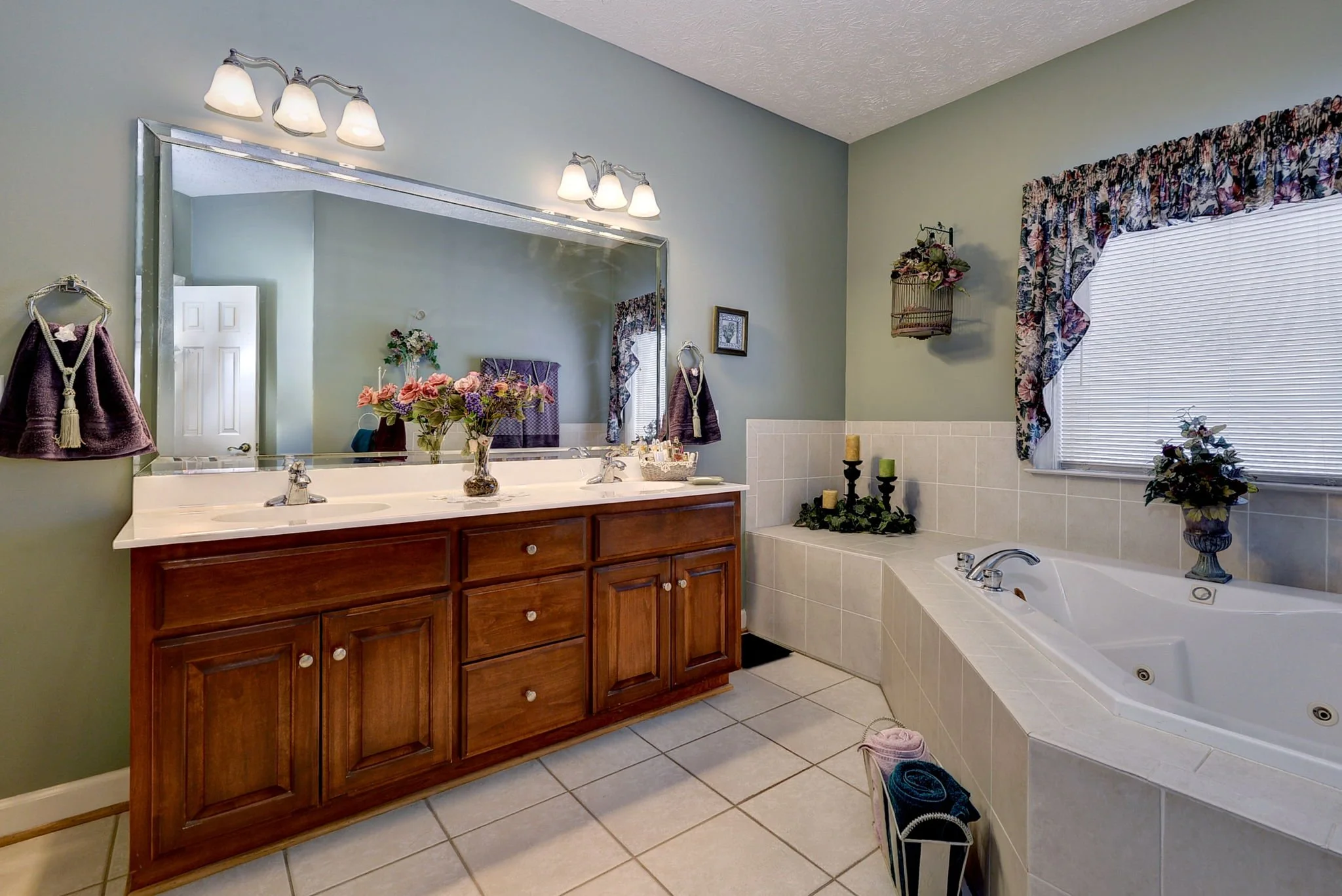 Bathroom with dual sinks, large mirror, wooden vanity, decorative flowers, candles, and a bathtub with plants, window, and floral valance.