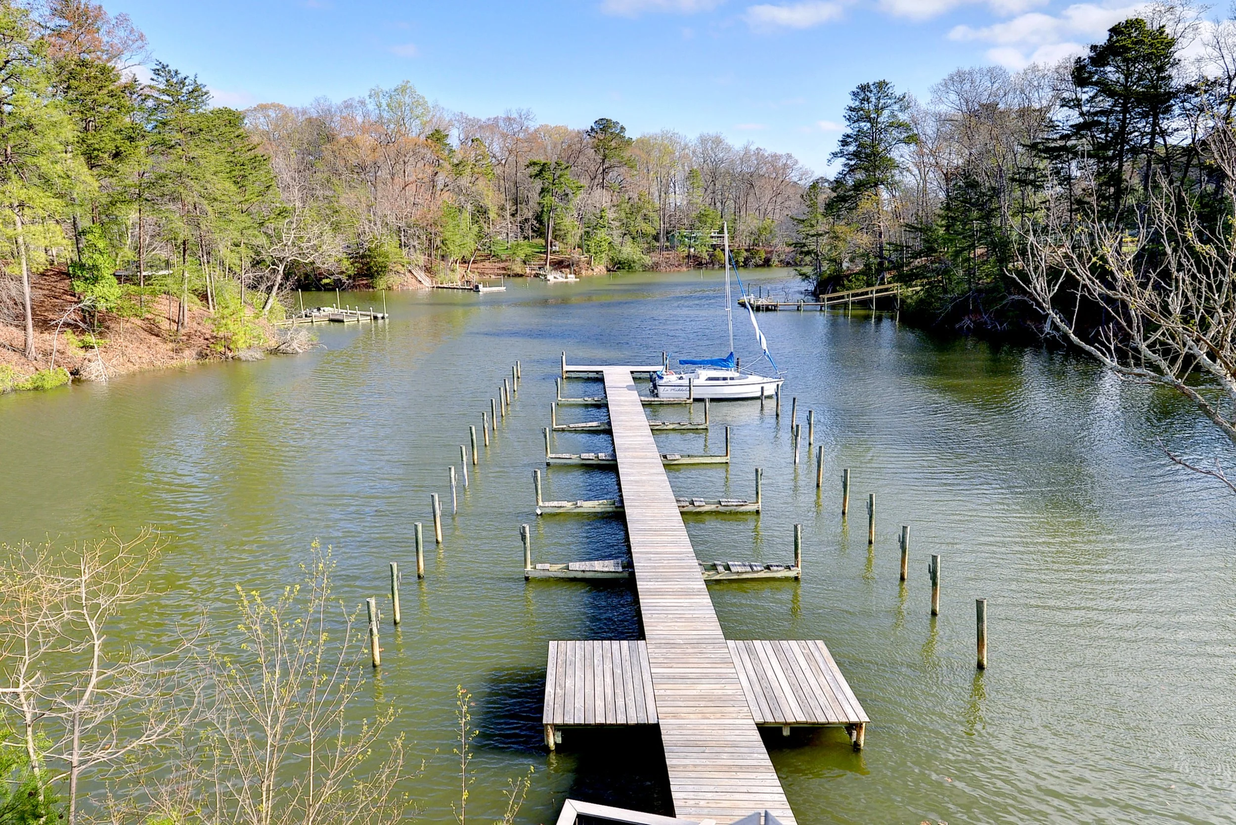 A wooden pier extending into a calm river with a sailboat docked at the end, surrounded by trees and a blue sky.