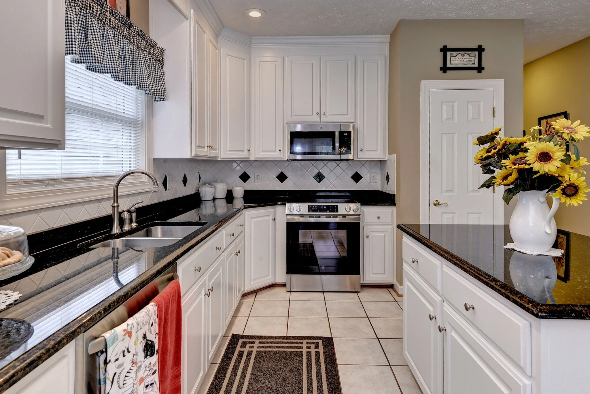 Kitchen with white cabinets, black granite countertops, a window with blinds and a checkered valance, stainless steel appliances including a microwave and oven, and a vase of sunflowers on the counter.