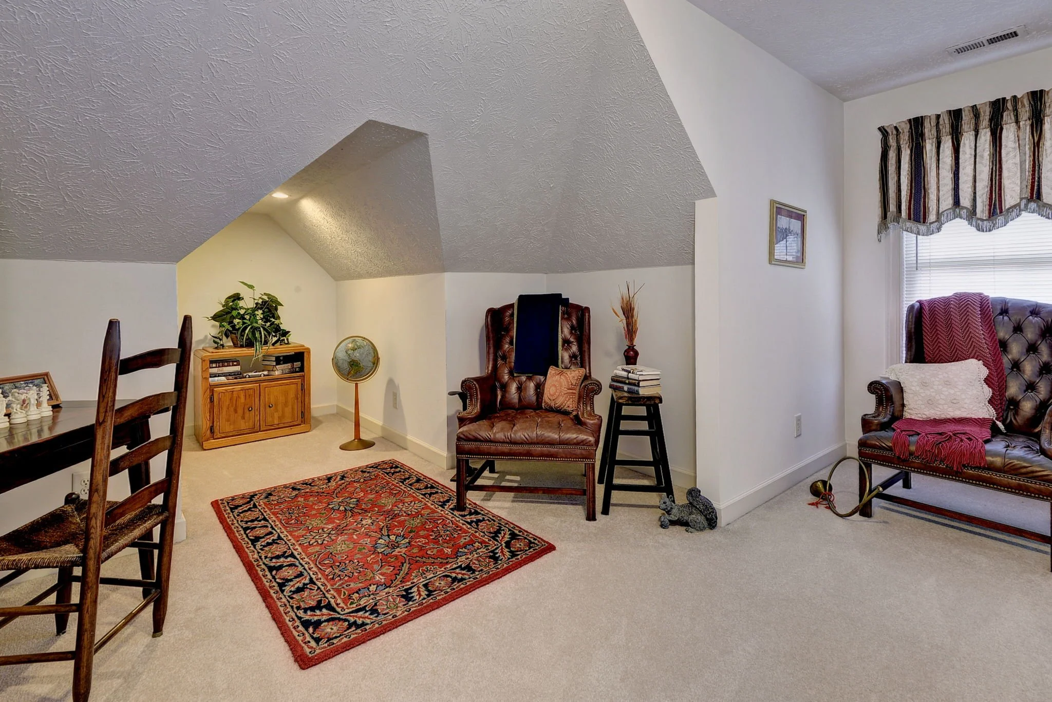 Living room area with sloped ceiling, a wooden dining table with chairs, a small wooden cabinet with plant, a world globe, an armchair with a pillow, a stack of books and decorative items, a window with striped curtains, and a second armchair with a 