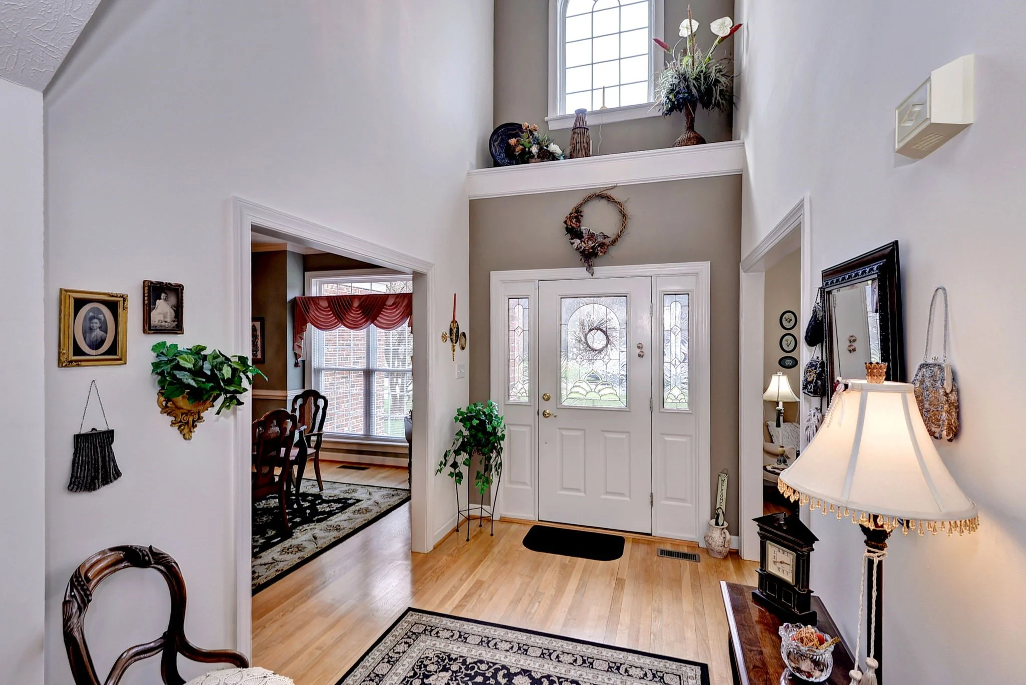 Entrance of a house with a white front door, decorative glass panels, a wreath, and a small black mat. Inside, there are houseplants, wall decorations, a standing lamp with a fringed shade, and a hallway leading to a dining room with a large window a