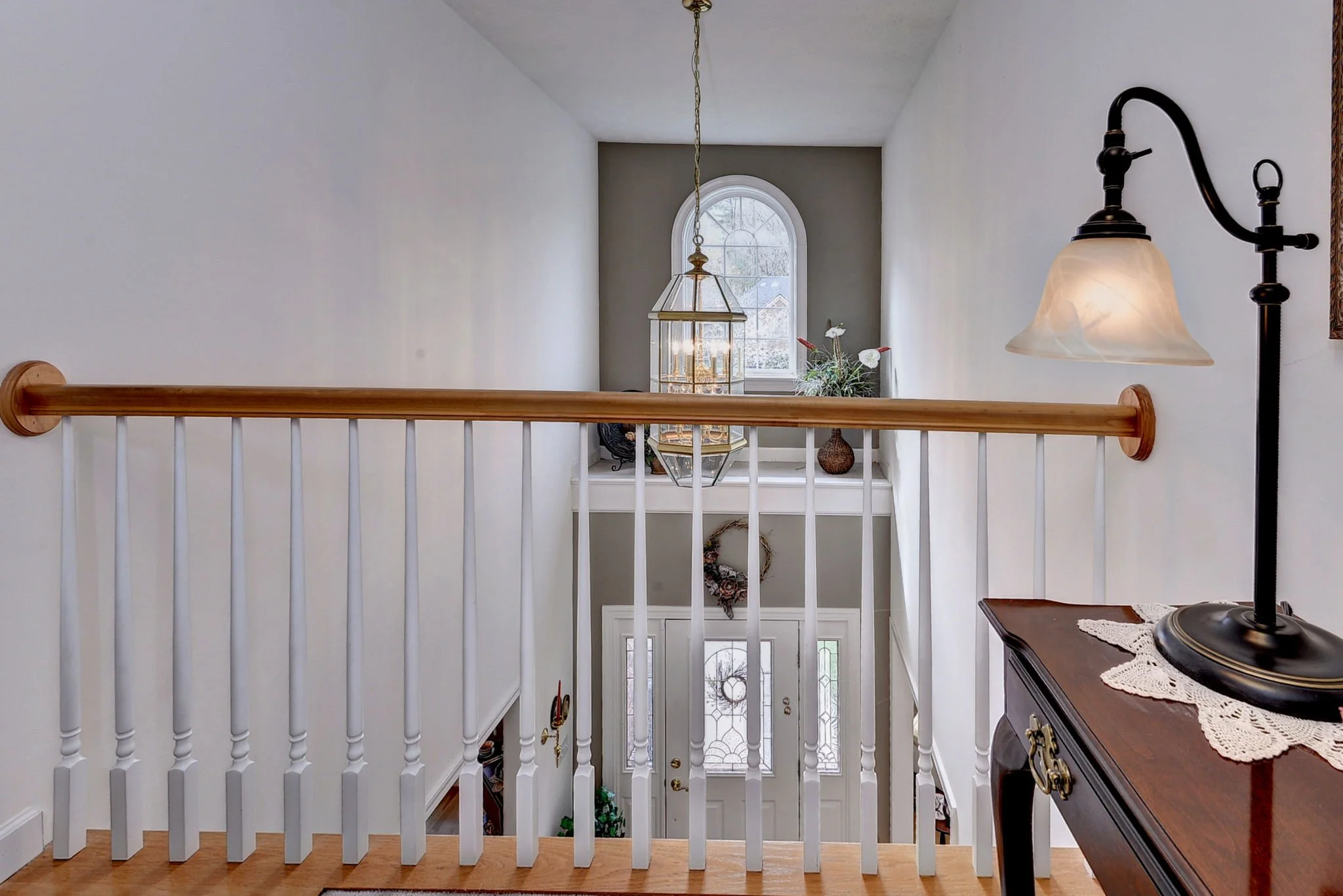 Interior view of a home's staircase landing with a white railing, a small table with a lace cloth and a lamp, and a window with a decorative wreath hanging below it, leading to the front door.
