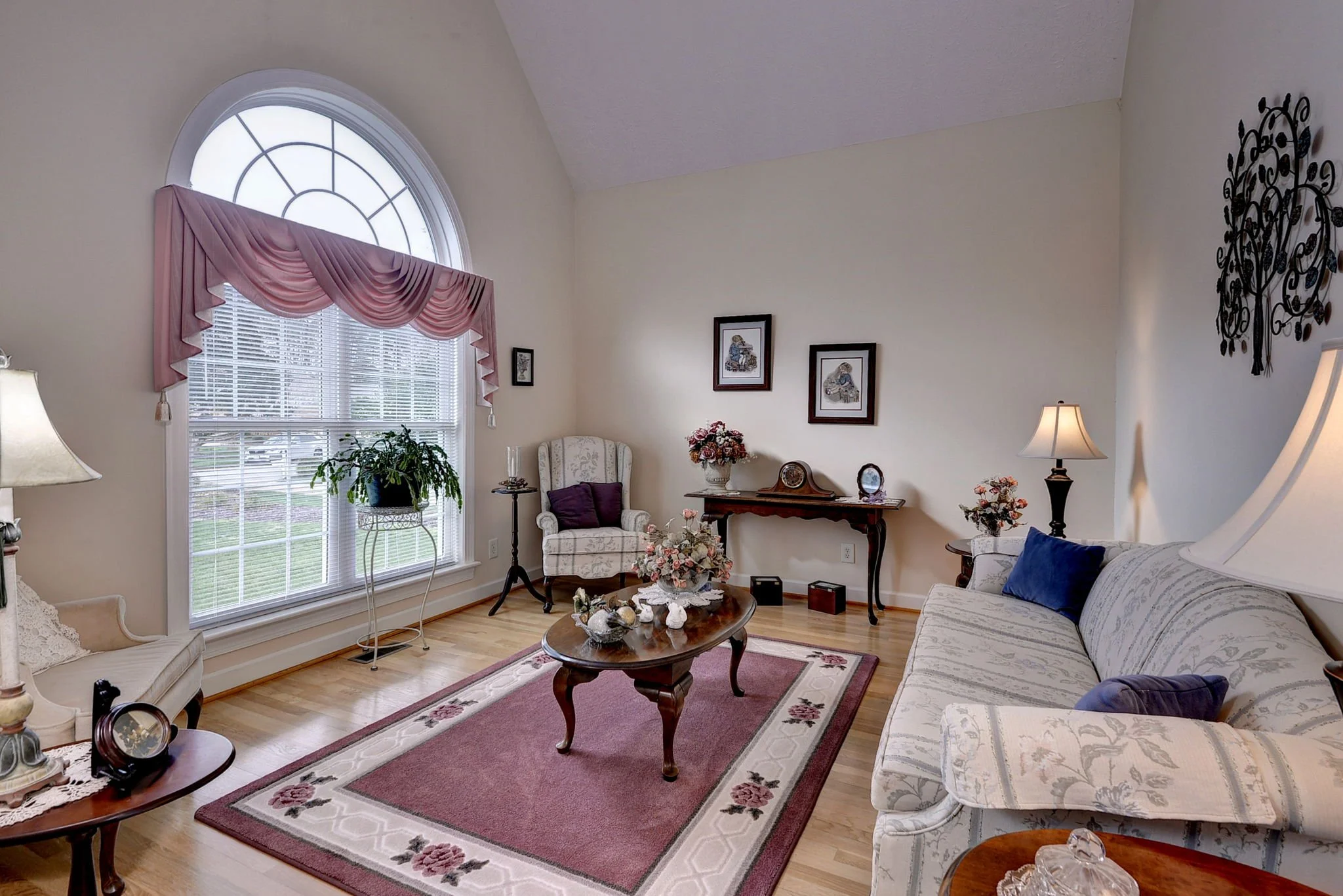 Living room with large arched window, pink valance, white sofa and armchair, floral decor, wooden coffee table, area rug, and wall art.