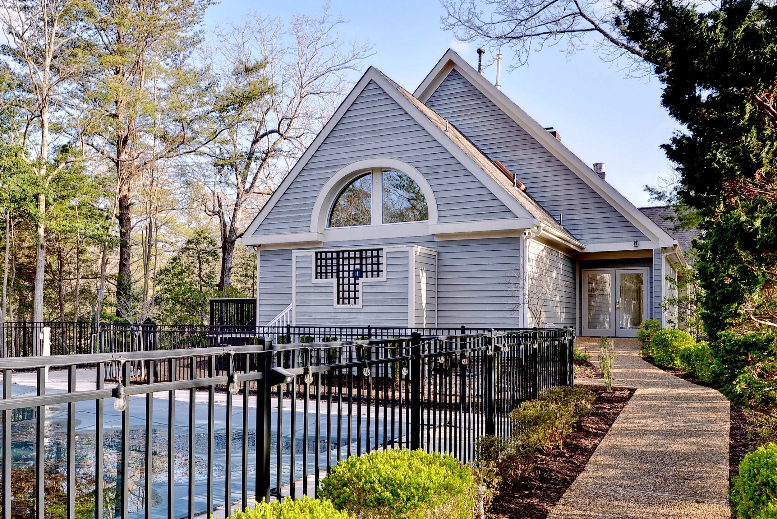 A light gray house with an arched window, surrounded by trees, with a paved walkway leading to a glass door, and a fenced swimming pool in the front yard.