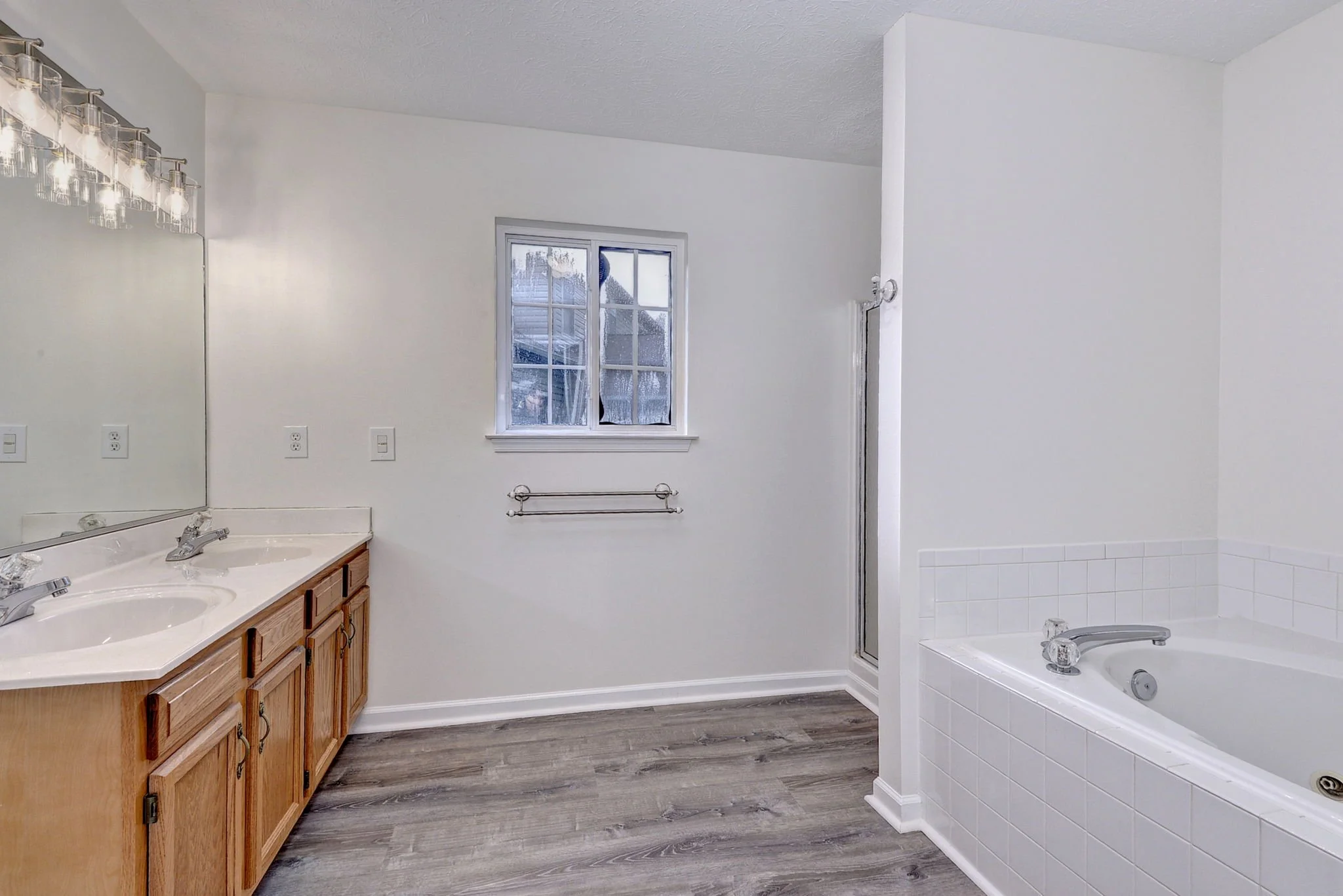Bathroom with white walls, double glass sink vanity with wooden cabinets, small window, and part of a bathtub with white tiles.