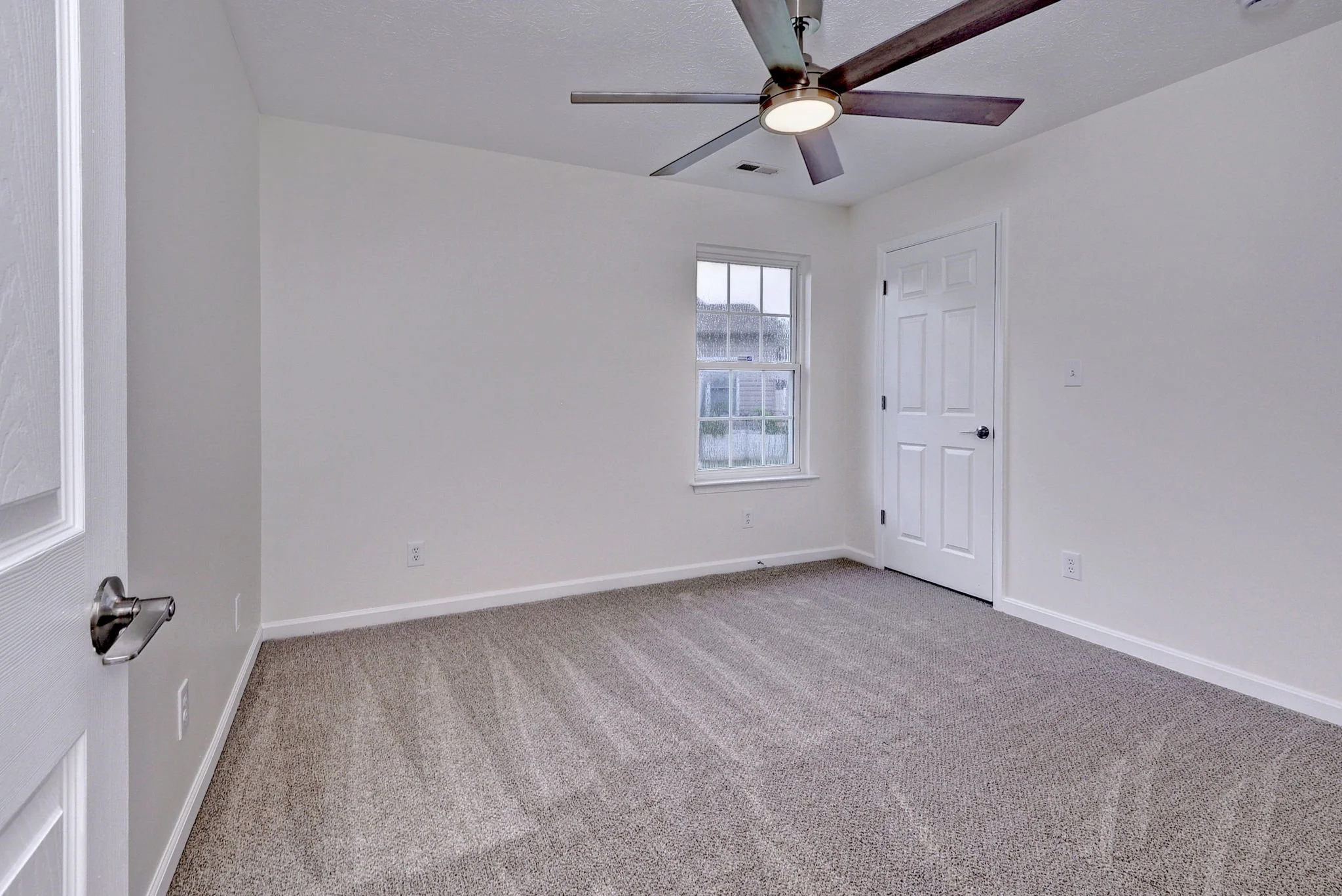 An empty room with white walls, beige carpet, a window with a view outside, a white door, and a ceiling fan with dark wooden blades.