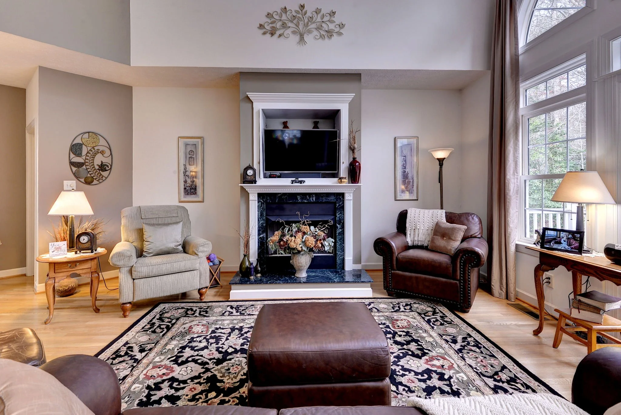 Living room with beige and brown furniture, a fireplace with a black marble surround, a wall-mounted TV above the fireplace, and large windows with beige curtains.