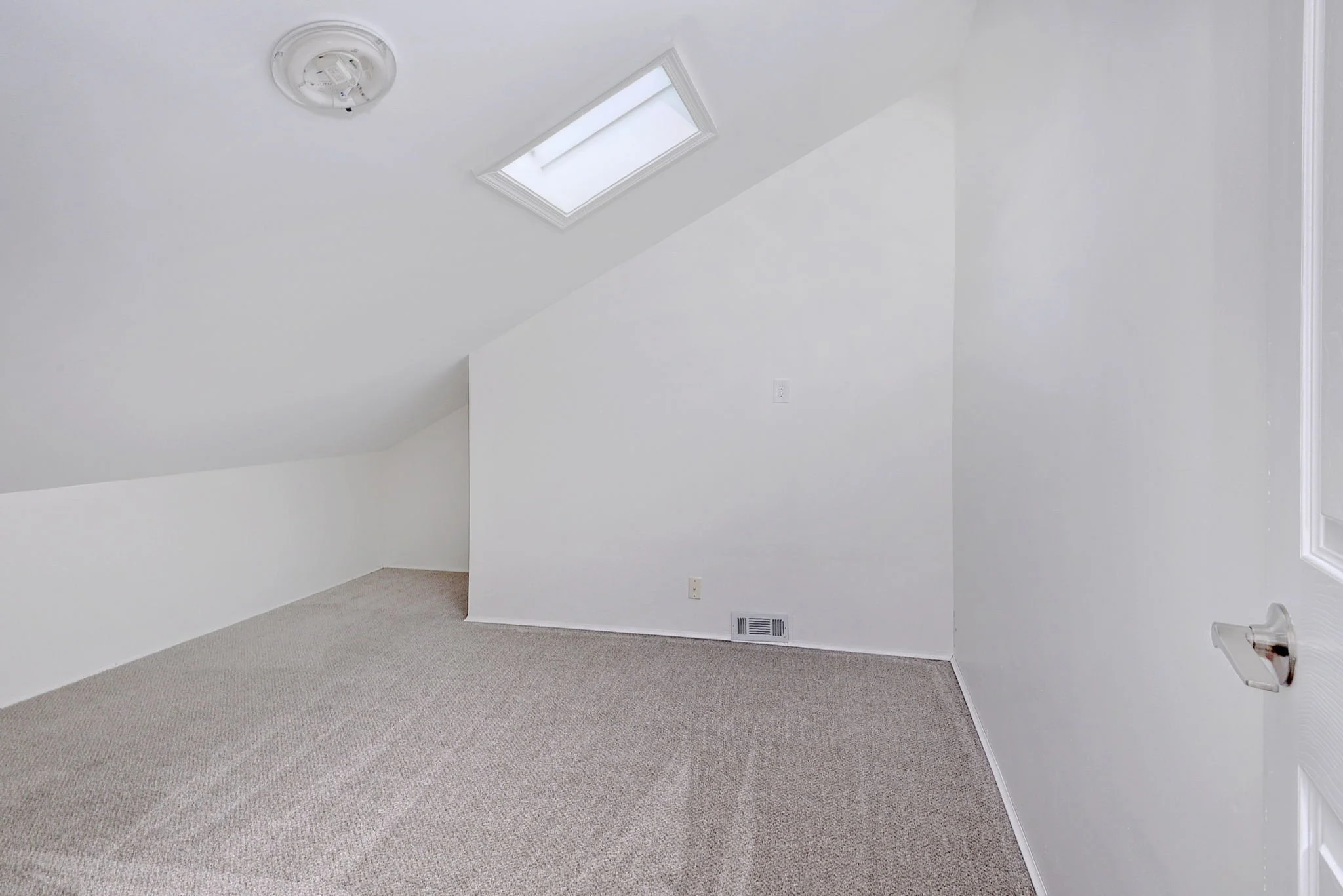 A white attic room with sloped ceiling, carpeted floor, a ceiling light fixture, a skylight window, and a wall vent.