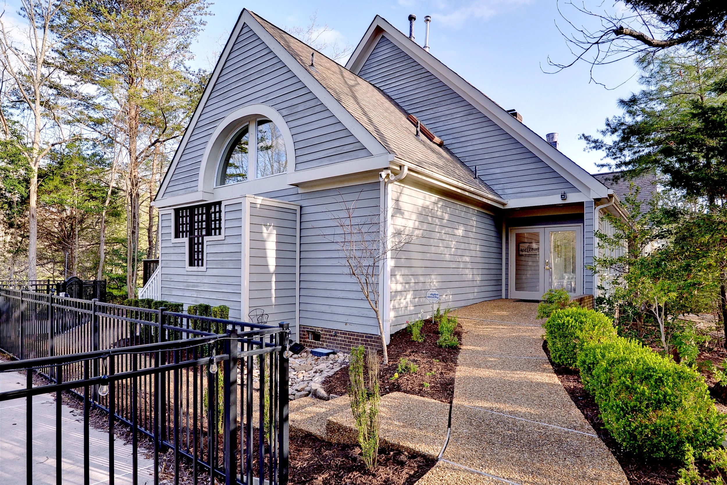 A house with light blue siding, a steep gable roof, and a large arched window. There is a pathway leading to the front door, surrounded by green shrubs and trees in a landscaped yard.