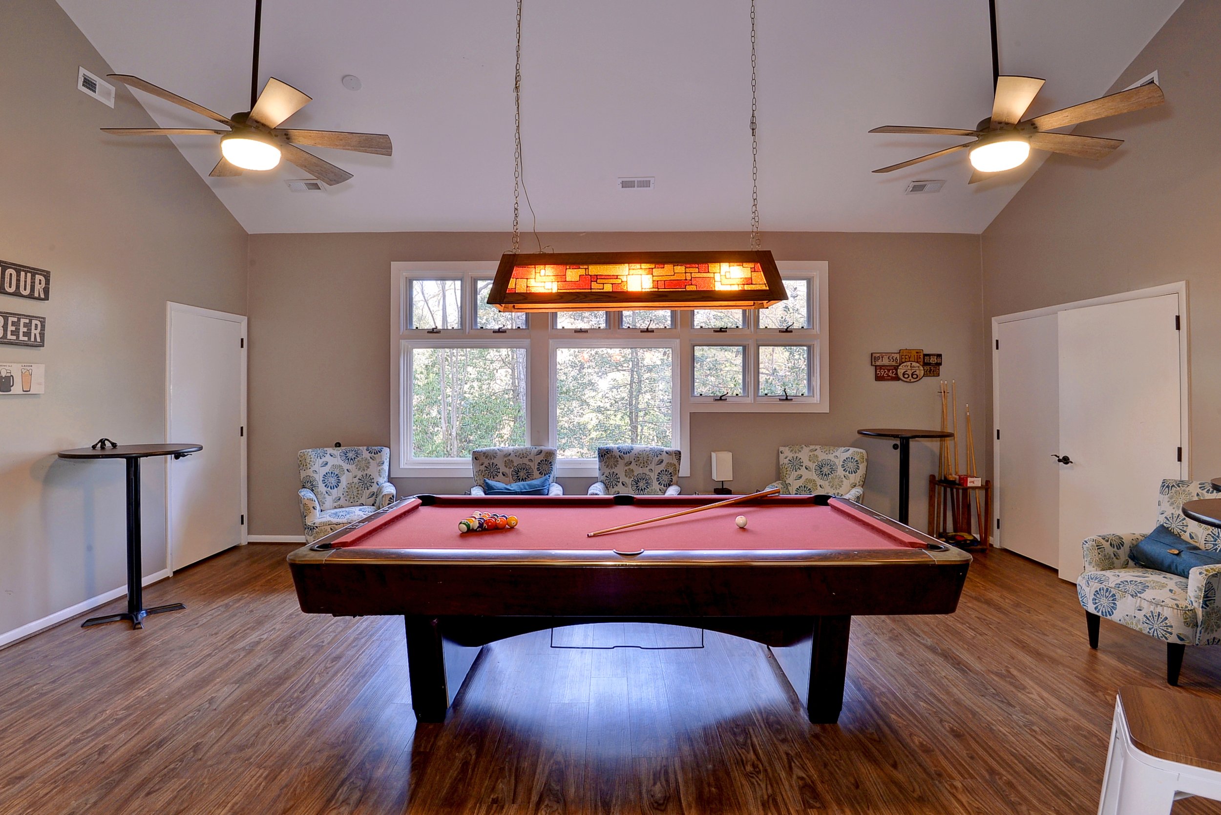 A living room with a red pool table, patterned armchairs, ceiling fans, and window views of trees outside.