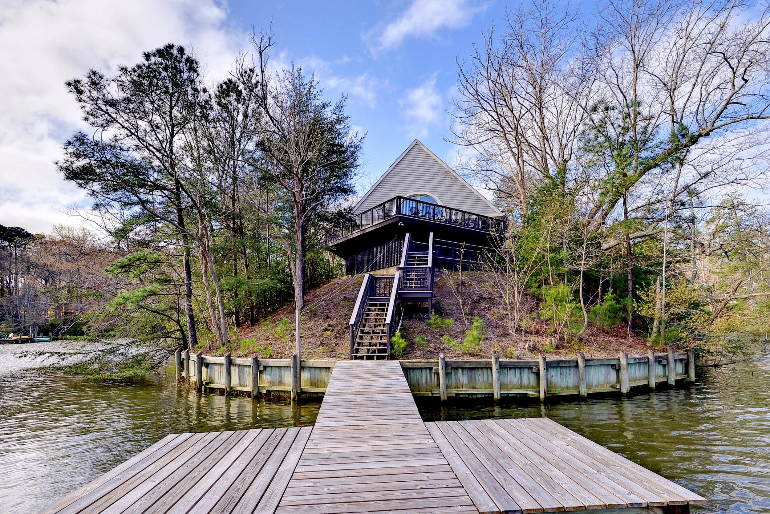 A house on top of a hill with a porch, seen from a dock over a body of water, with trees surrounding the house and a partly cloudy sky.