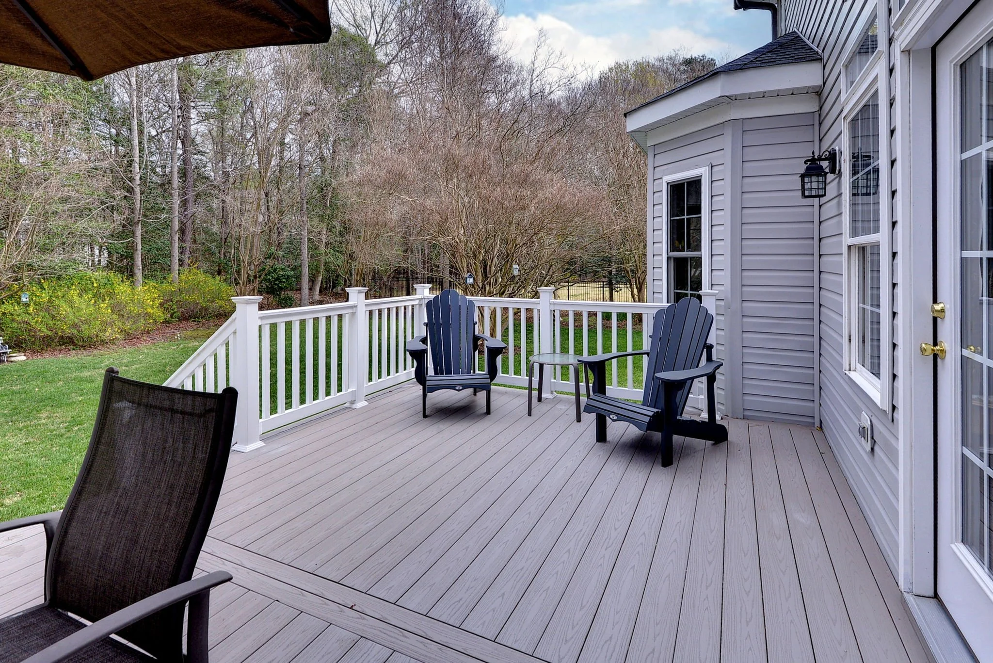 A wooden deck with two black Adirondack chairs, a small round table, and a patio chair with a wooden overhang. The deck is surrounded by a white railing and overlooks a yard with green grass and trees.