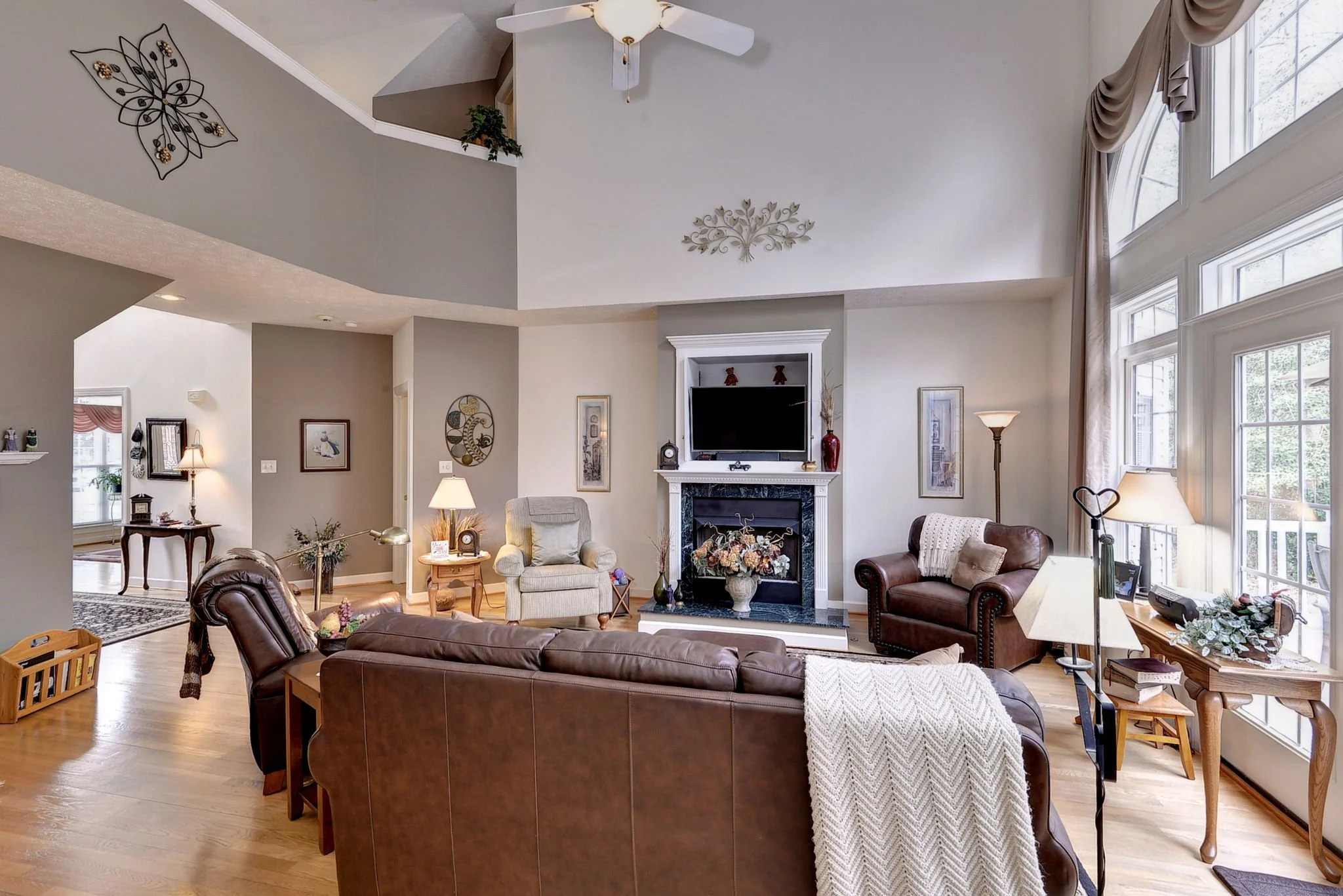 Spacious living room with large windows, leather and fabric armchairs, a white fireplace with a TV, a ceiling fan, and decorative wall art.