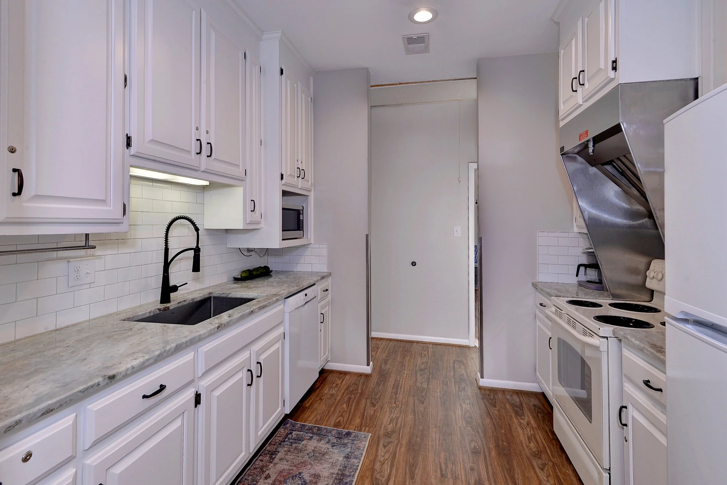 Empty kitchen with white cabinets, marble countertops, and white tiled backsplash. Contains a black faucet, microwave, and electric stove with vent hood. Wooden floor and a small rug in front of the sink.