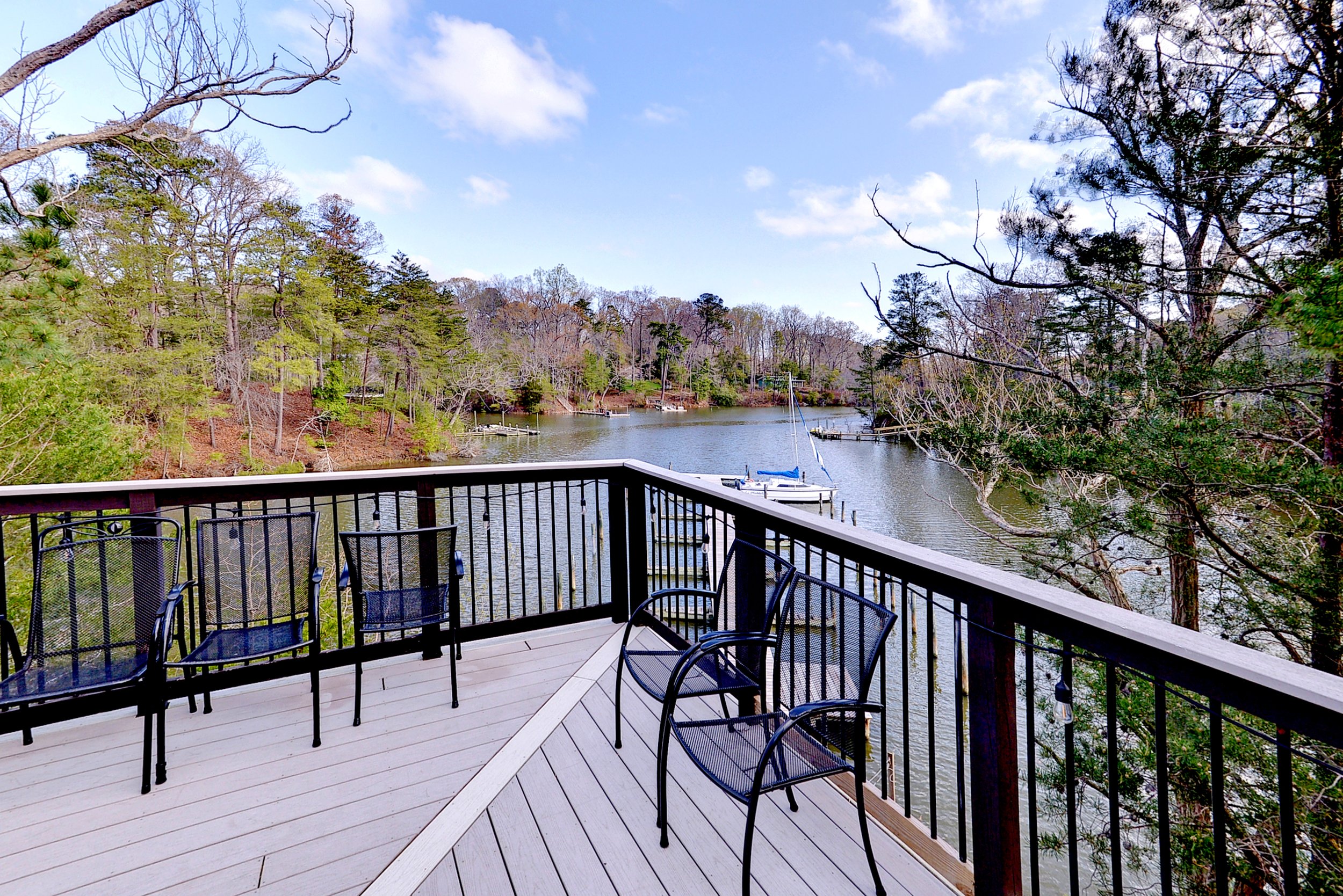 A wooden deck with black metal chairs overlooking a calm river, surrounded by trees with some bare branches and others with green foliage, under a partly cloudy sky.