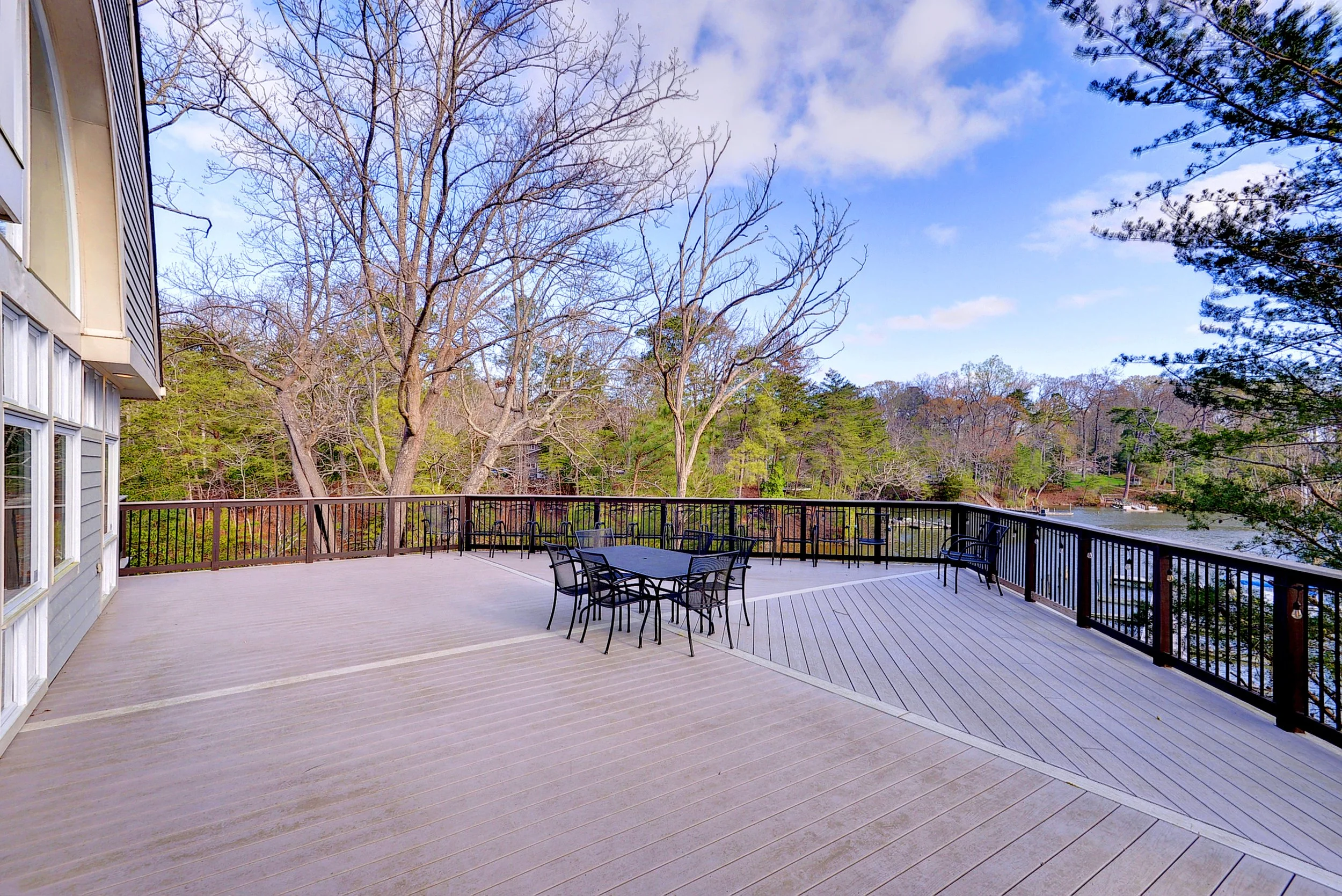 Large wooden deck with metal patio furniture overlooking a lake, surrounded by trees, with a house on the left side and a partly cloudy sky.