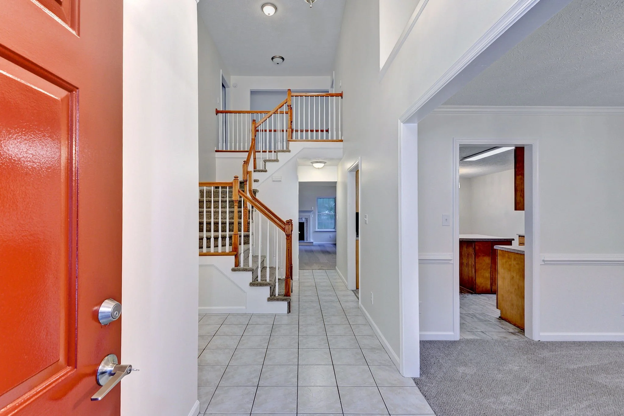 Interior view of a house entryway with a red front door, tiled floor, staircase with wood railing, and white walls.