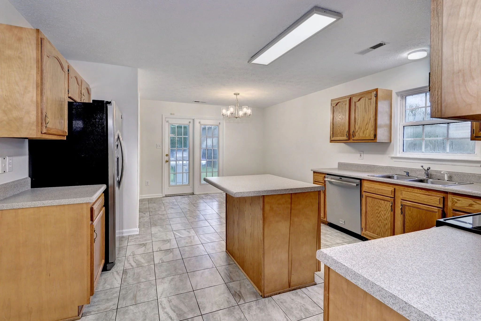 A kitchen with wooden cabinets, a black refrigerator, tiled floor, and a countertop with a sink. There is a hanging chandelier and double doors leading outside, with windows on the right side.