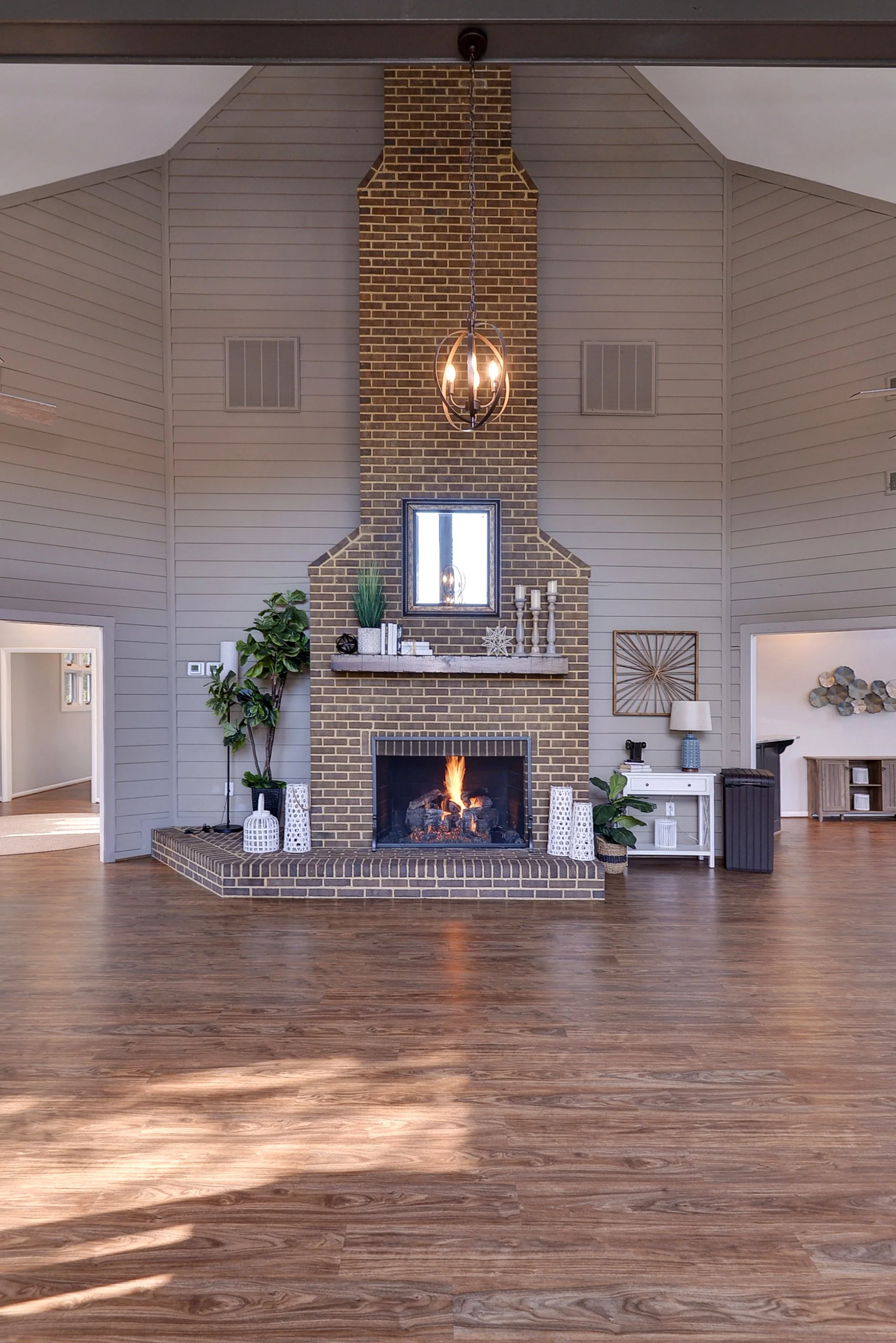 Living room with brick fireplace, wooden mantel, and decor including plants, candles, and mirror, with hardwood floors and vaulted ceilings.