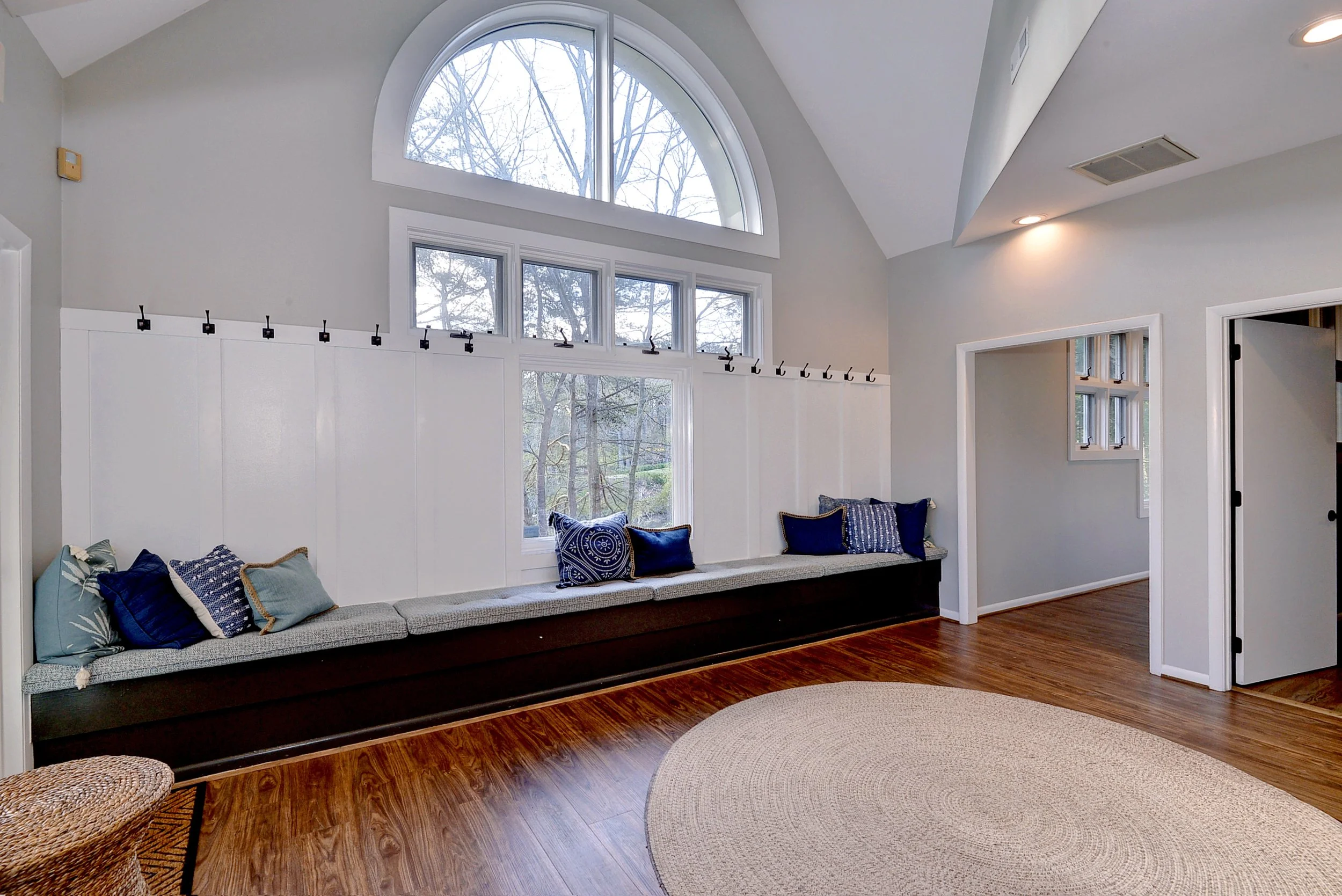 Interior of a room with a large arched window and a window seat with blue and patterned throw pillows, white paneled walls, and wooden flooring.