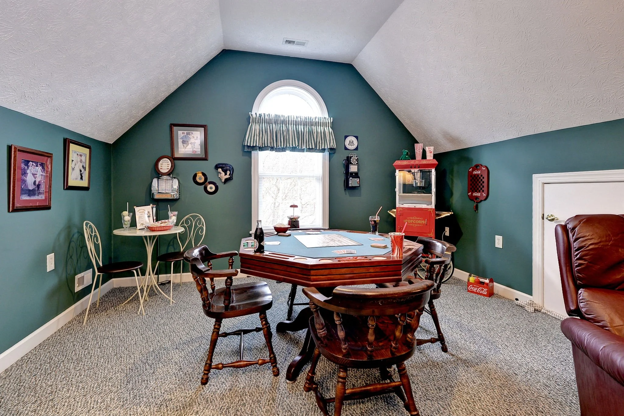 A cozy room with a green accent wall, vaulted ceiling, and a window with striped valance. Features include a round metal table with two chairs, a game table with four wooden chairs, and a popcorn machine. Decor includes framed pictures, vintage items