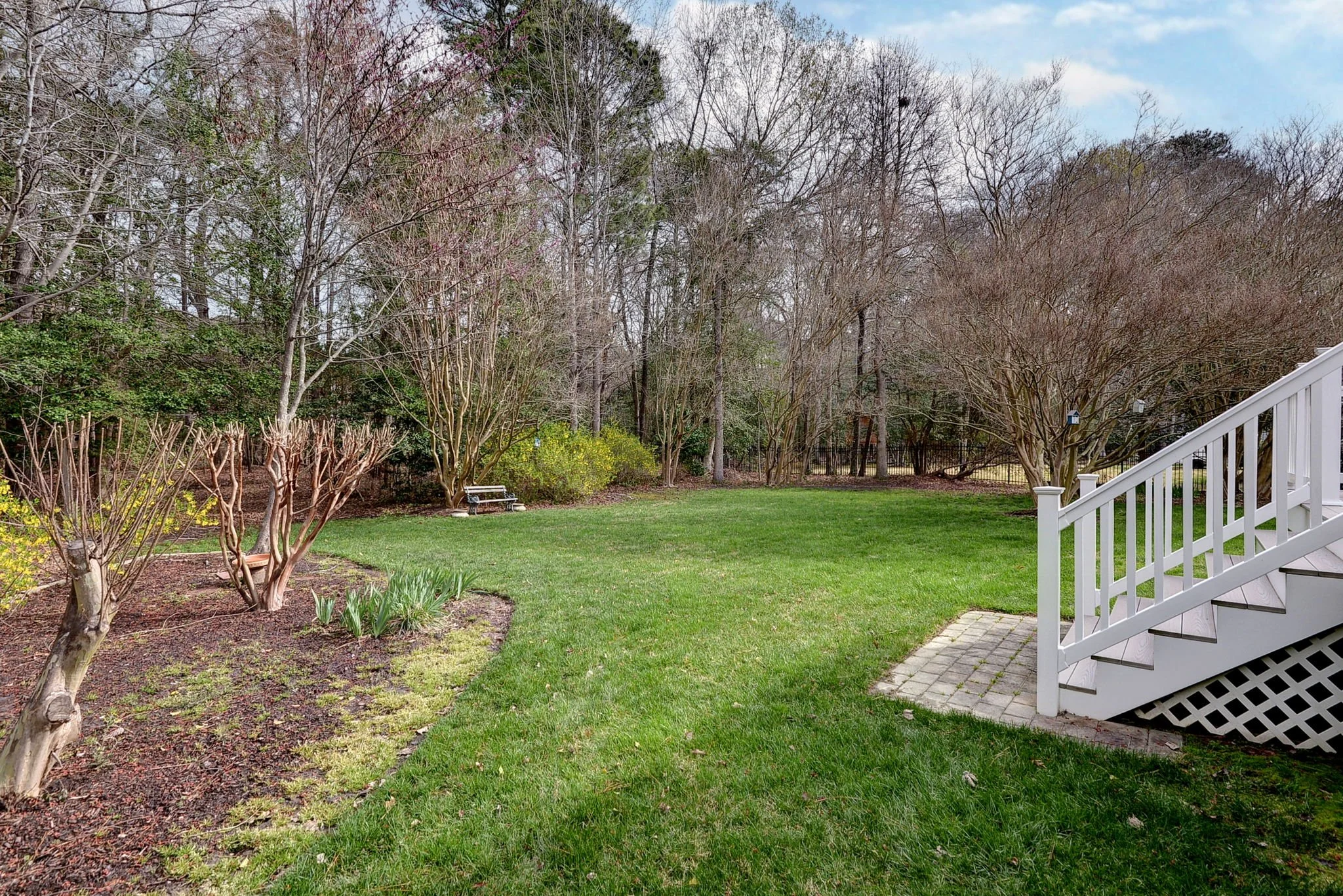 View of a backyard garden with a lush green lawn, trees with sparse leaves, a white garden bench, and a staircase leading from a deck or porch on the right side. Overcast sky in the background.