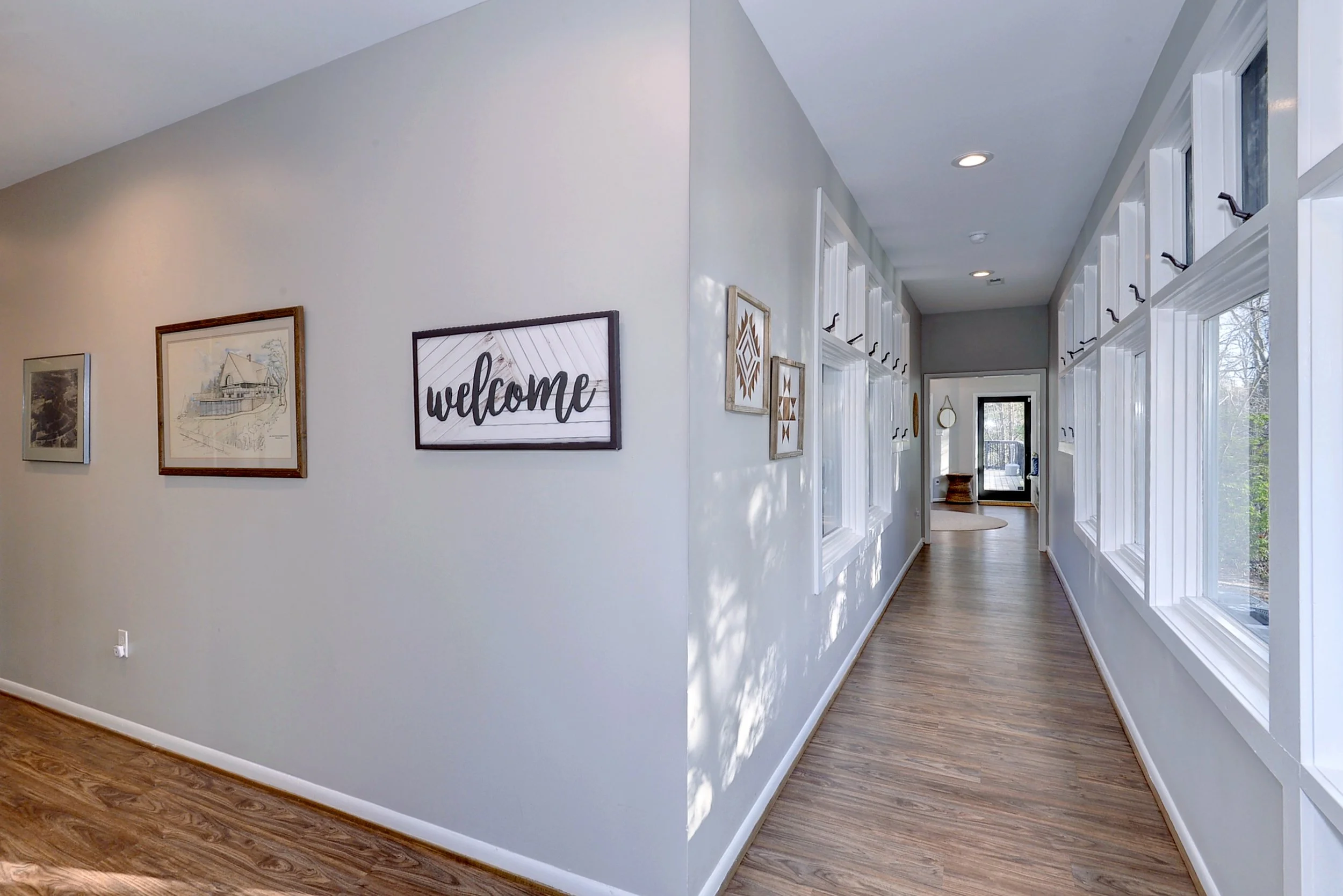 Bright hallway with large windows on the right side, white walls, wooden floors, and wall decor including framed pictures and a 'welcome' sign.