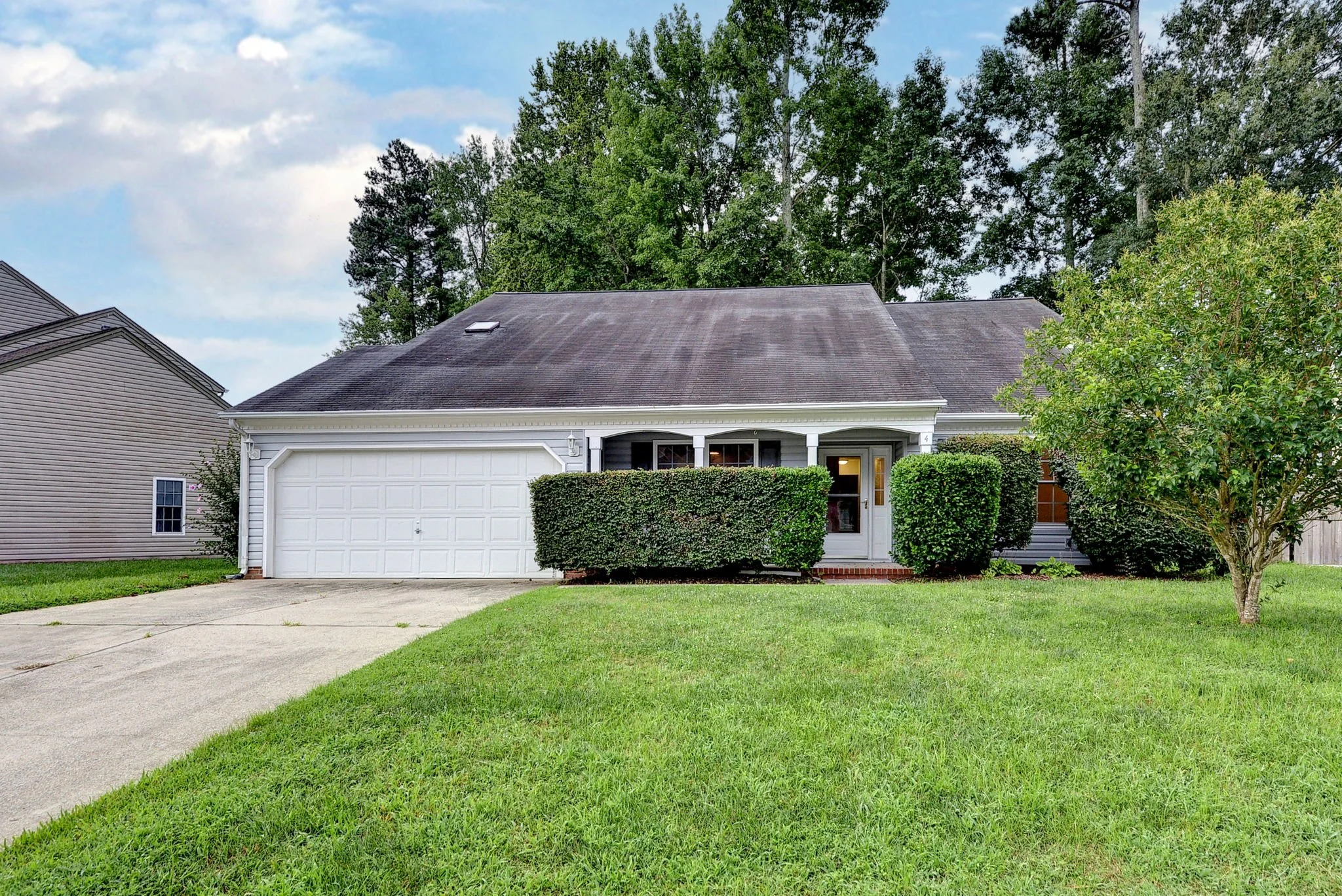 A suburban house with a white garage door, a front door with a window, and well-maintained green bushes and lawn. There are trees in the background and a paved driveway.