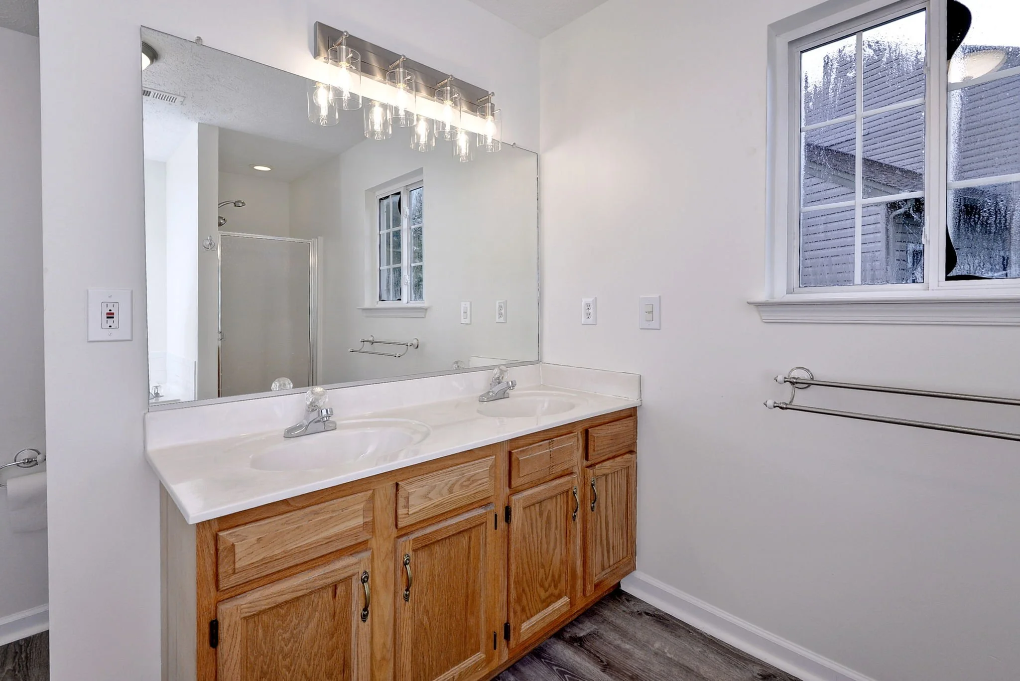 Bathroom with white walls, a large mirror, two sinks with a wooden vanity, a window showing outdoor rain, and a chrome towel rack.
