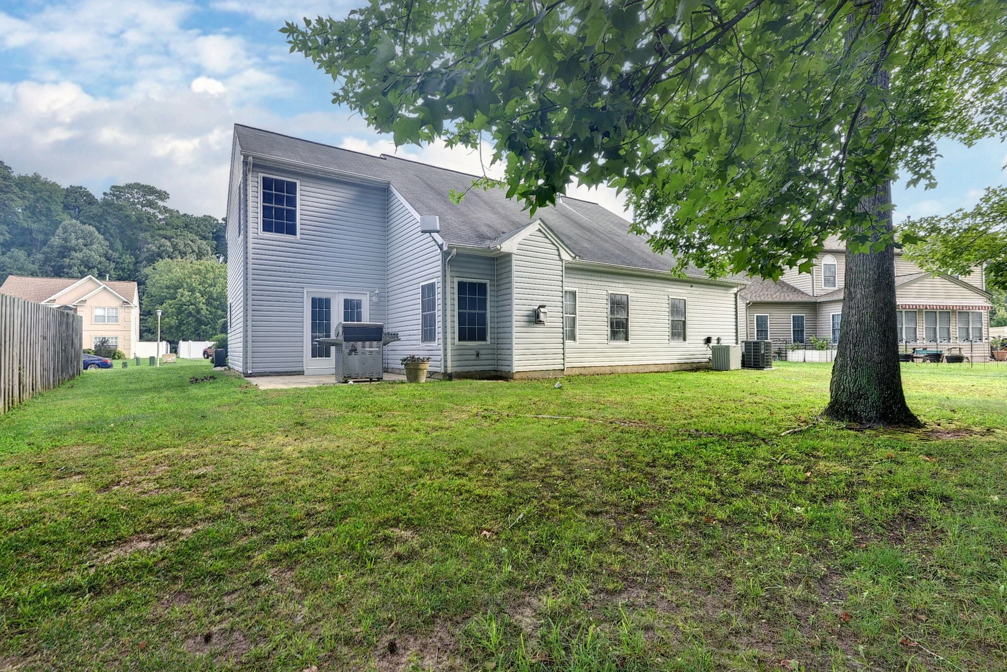 Backyard grassy area with large tree, white house with vinyl siding, several windows, patio, air conditioning units, and a fenced side yard in a suburban neighborhood.
