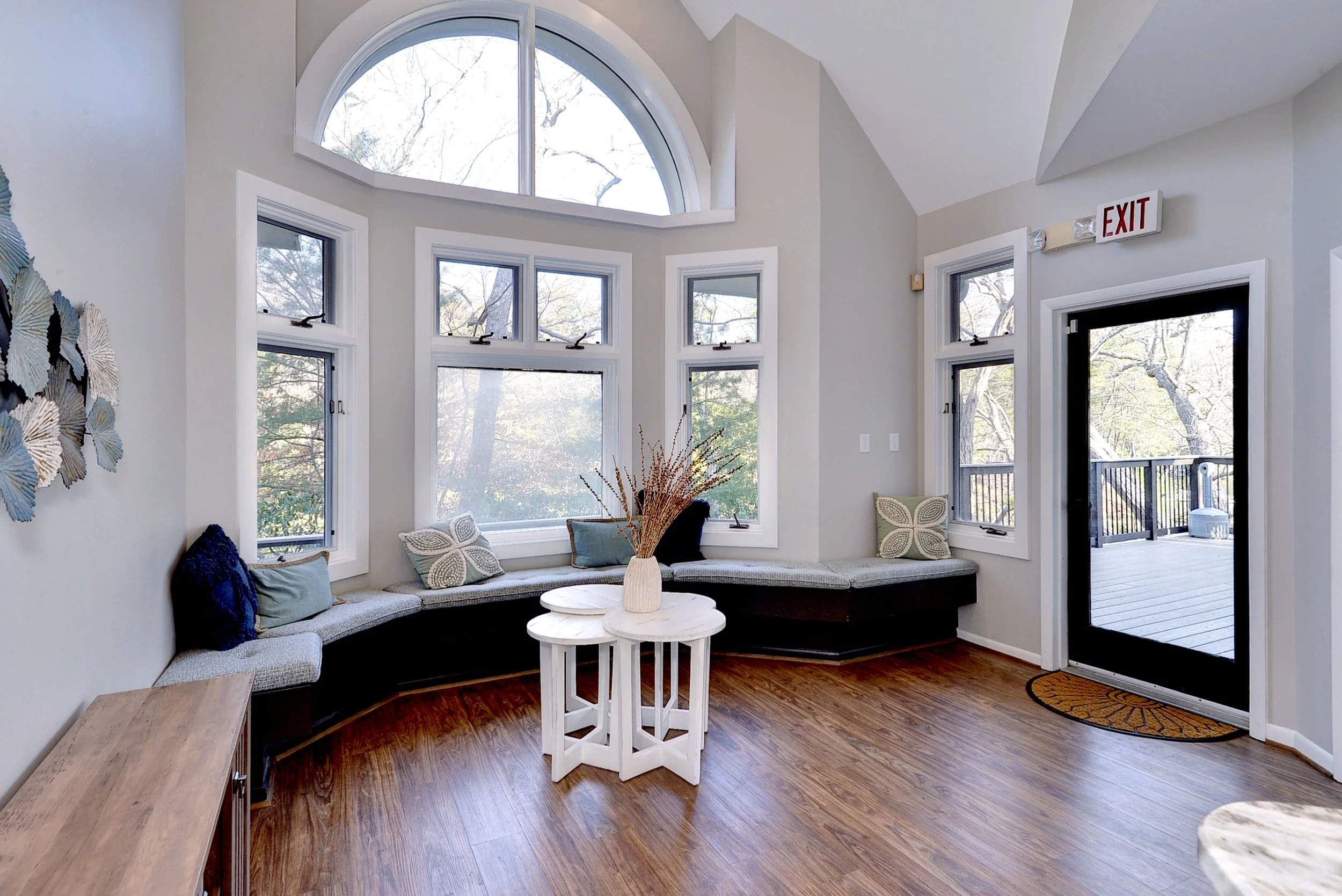 A cozy sunroom featuring a curved bench with pillows beneath large bay windows and a glass door leading to an outdoor balcony. A white side table with a vase and dried flowers sits in the center. Light-colored walls and hardwood floors complete the w