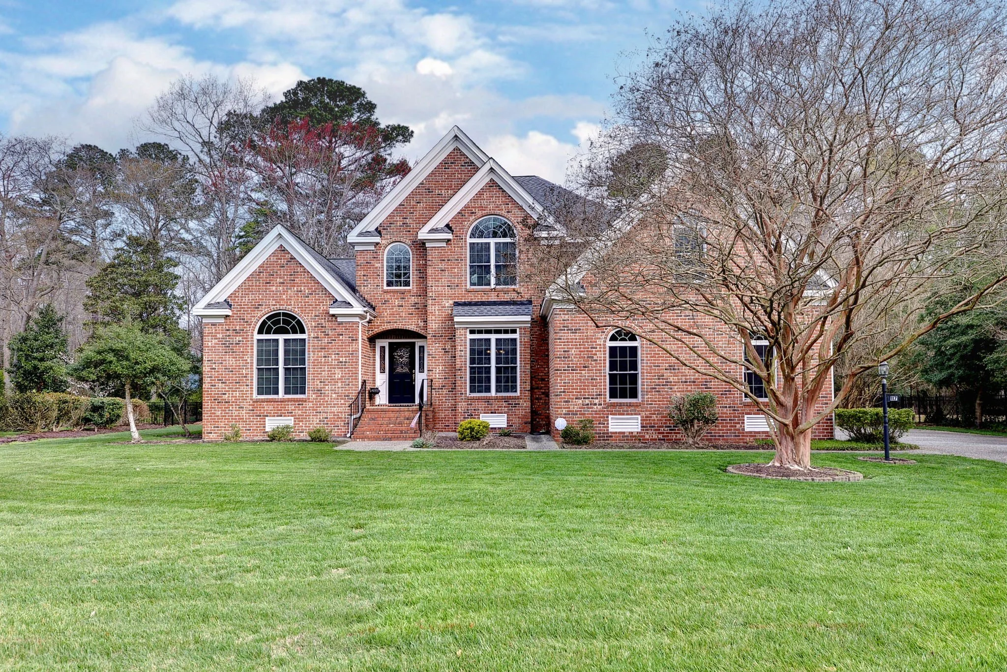 A large brick house with multiple gables and arched windows. The front yard has a well-maintained lawn, a leafless tree, small shrubs, and a driveway on the right side.