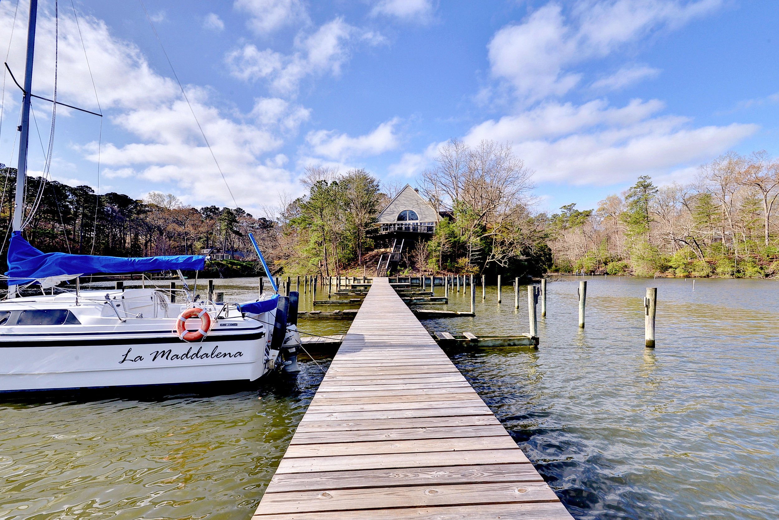 A wooden dock extending into a river with a sailboat named La Maddalena moored on the left side. The background features a house on a hill surrounded by trees, under a partly cloudy sky.