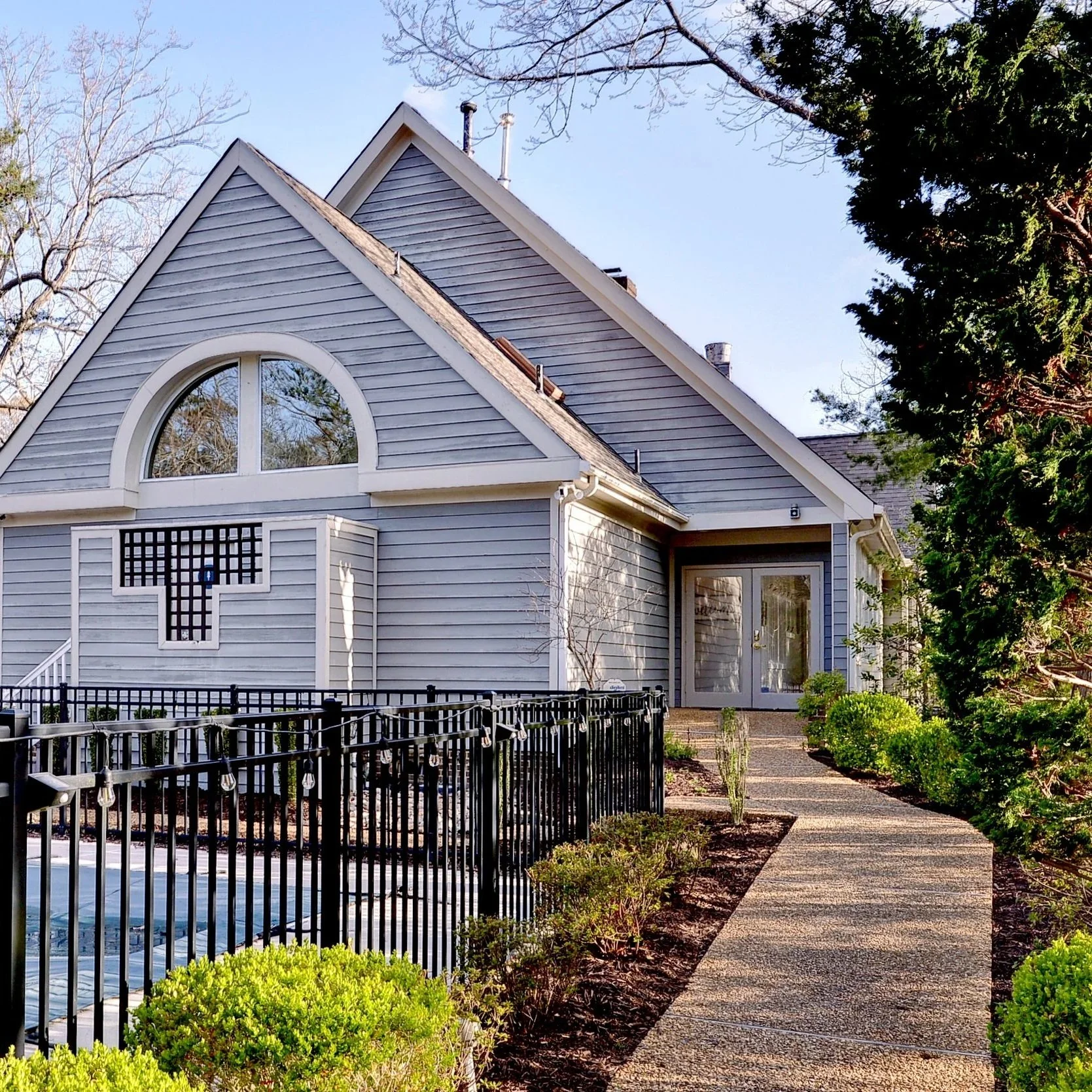 A white house with gray trim and a triangular roof, glass double doors, a walkway lined with bushes, and a black metal fence surrounding a pool.