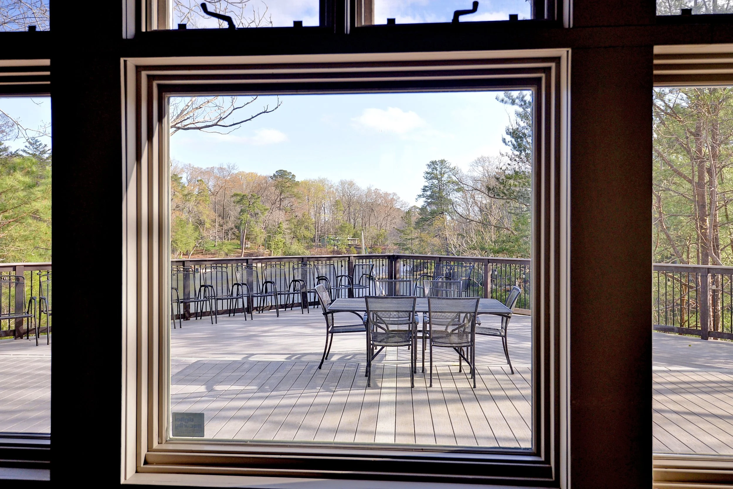 View of a wooden outdoor balcony with a round table and chairs, framed by a sliding glass door, overlooking a scenic landscape with trees and a blue sky.