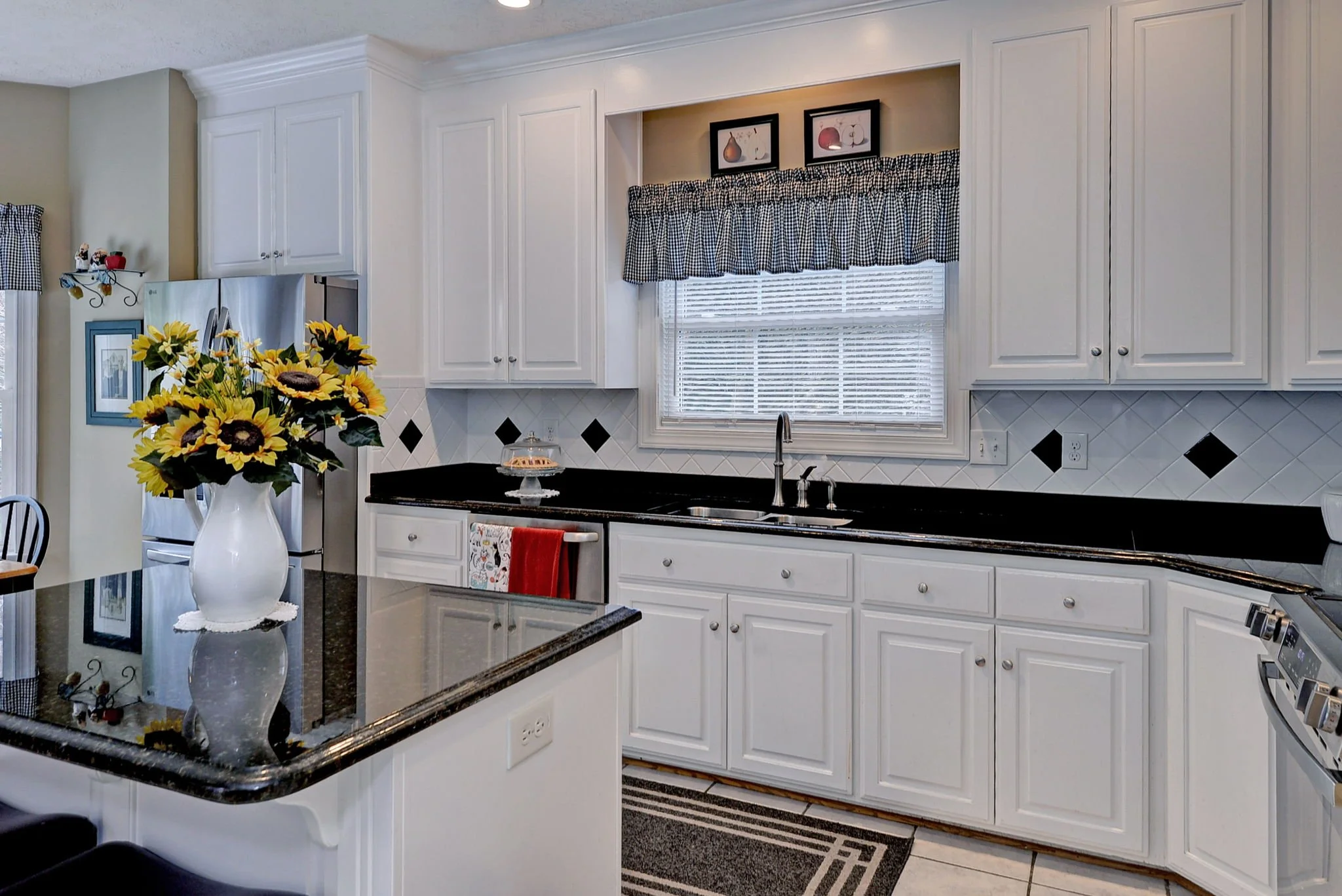 Kitchen with white cabinets, black countertops, a window with checkered valance, vase of sunflowers on island, stainless steel refrigerator, and black and white diamond tile backsplash.