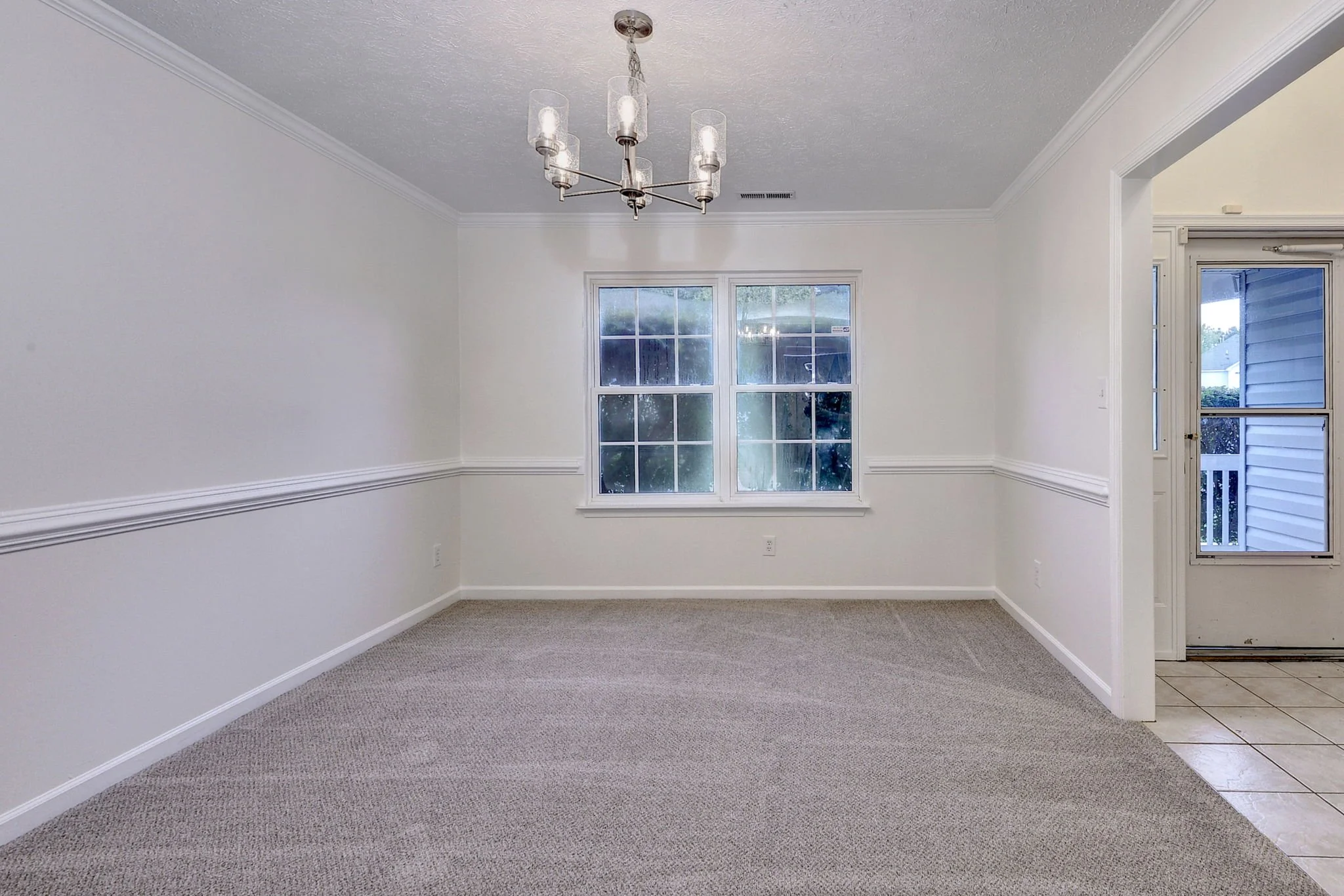 Empty living room with white walls, carpeted floor, large window, and a chandelier hanging from the ceiling near the window. Part of the adjacent kitchen with tiled floor is visible on the right.