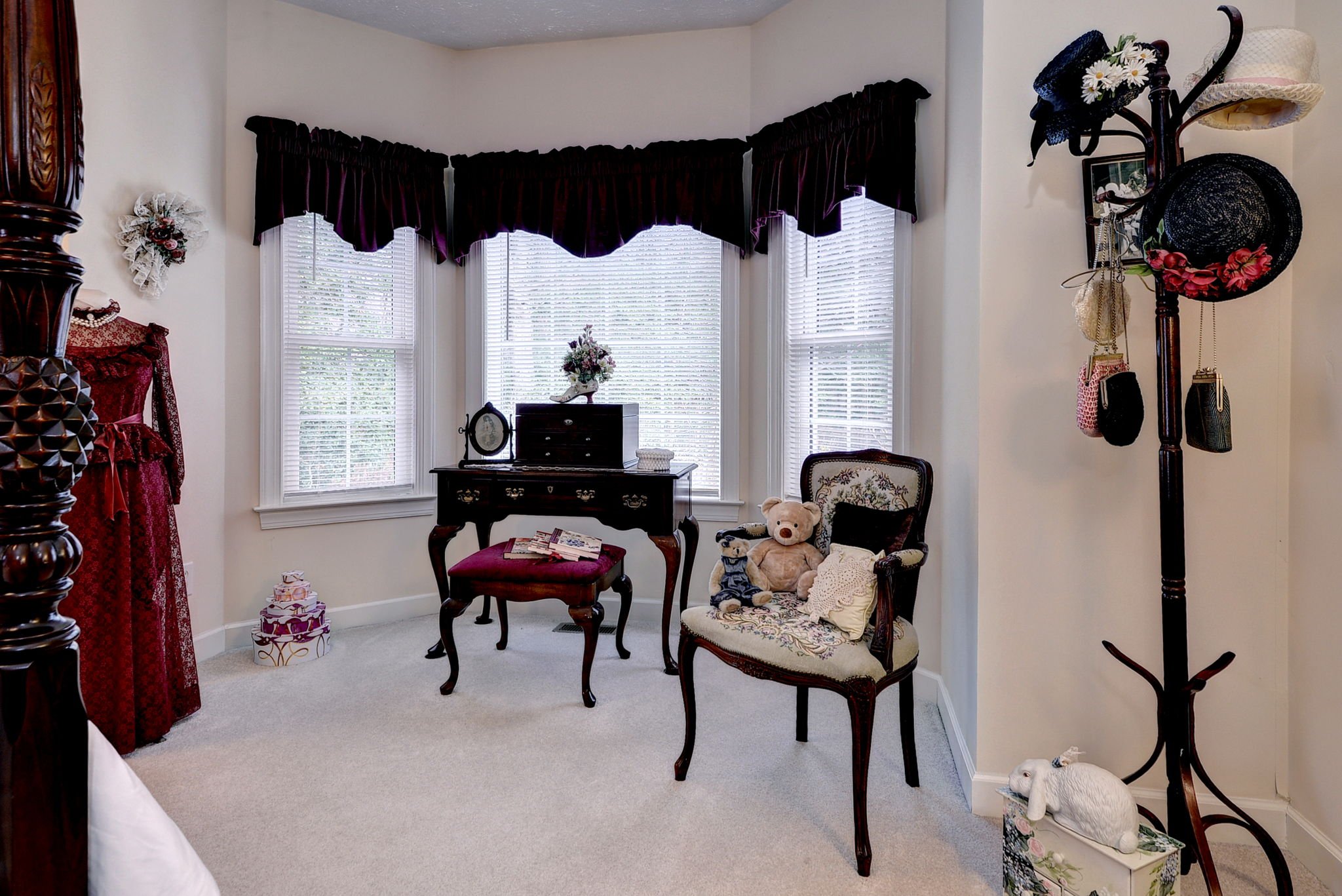 A cozy bedroom corner with a vintage vanity desk, a floral chair with teddy bear and doll, a coat rack with hats and purses, and a window with dark curtains and white blinds.